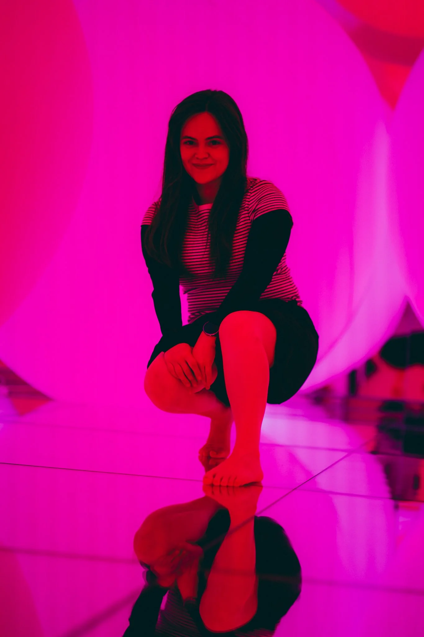 A woman with long dark hair, wearing a striped shirt and black sleeves, is crouching barefoot on a reflective floor in front of a pink-lit art installation or background.