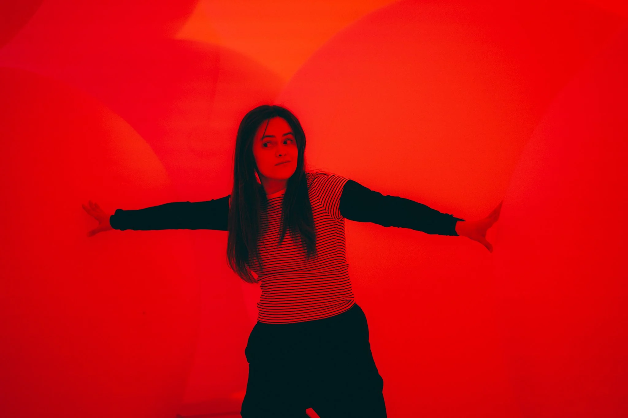A woman with long dark hair wearing a striped shirt and black sleeves extends her arms against a red glowing background.