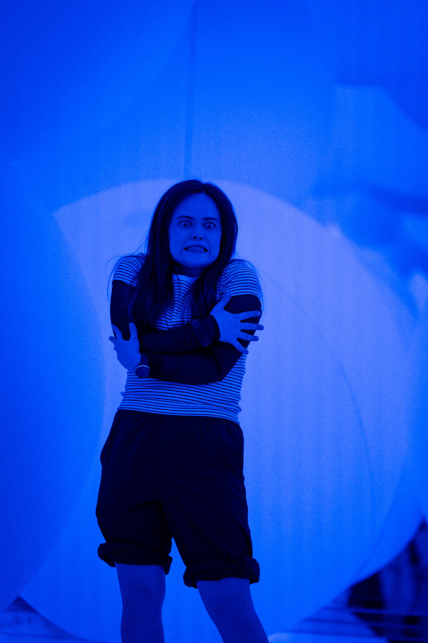 A woman with long dark hair, wearing a striped shirt and black shorts, stands with her arms crossed over her chest and a distressed or frightened expression on her face, illuminated in blue light.