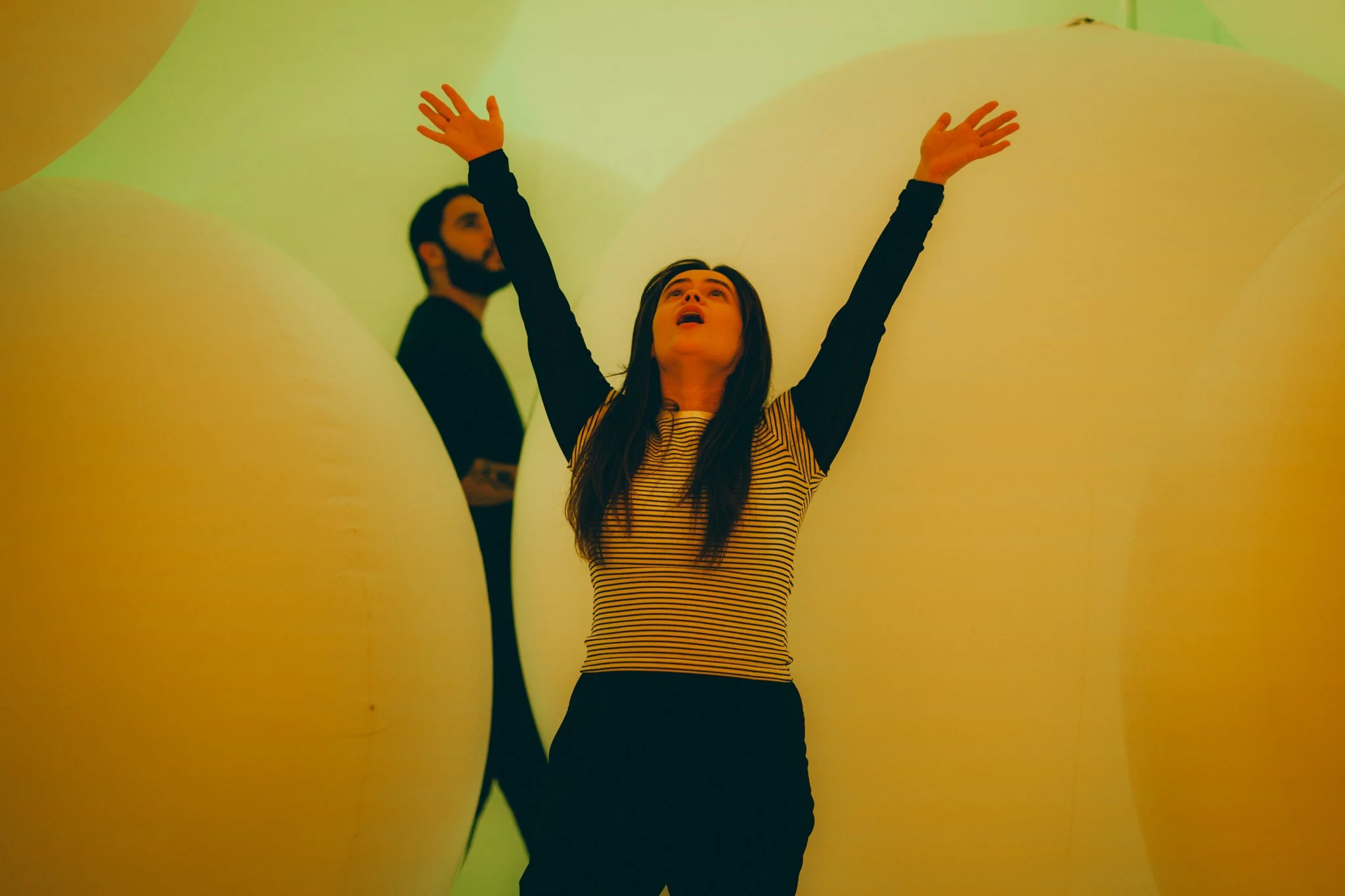A woman with long dark hair, wearing a striped shirt, raising her arms upward while standing between large yellow spherical objects in an art installation. A man with a beard, dressed in black, stands in the background.