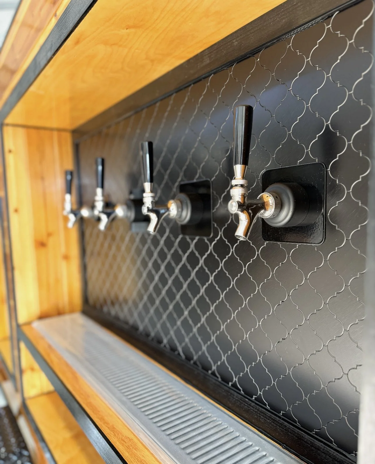 Four beer taps mounted on a black tiled wall above a metal drip tray with a wooden frame.