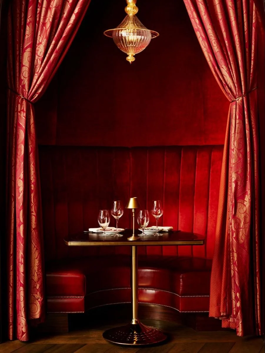 Intimate dining nook with red velvet walls, curtains, and banquette seating, set with a small table, four wine glasses, and a gold lamp, lit by a hanging chandelier.