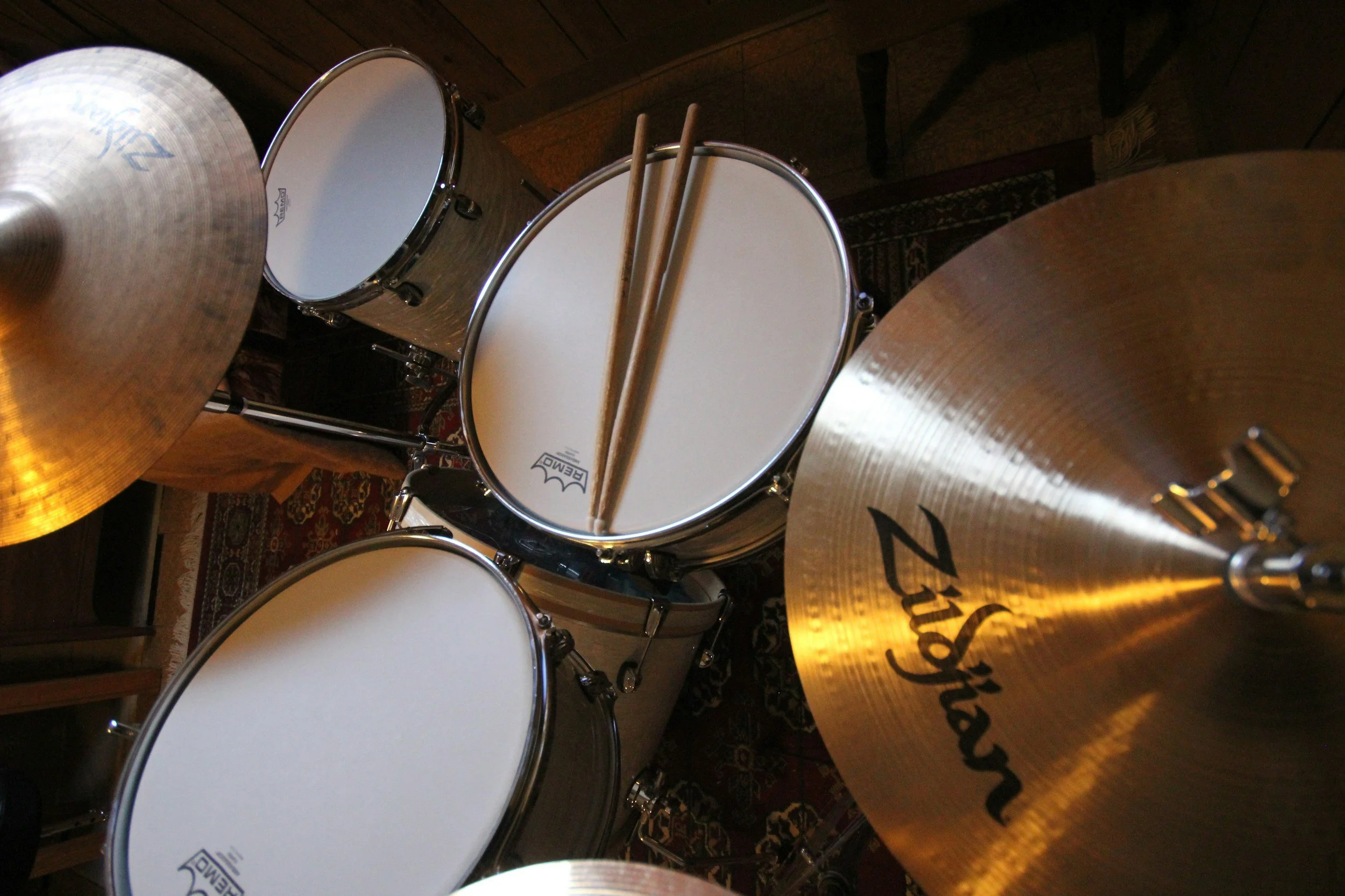 A drum set with cymbals, toms, and drumsticks on top, placed on a wooden floor with a patterned rug underneath.