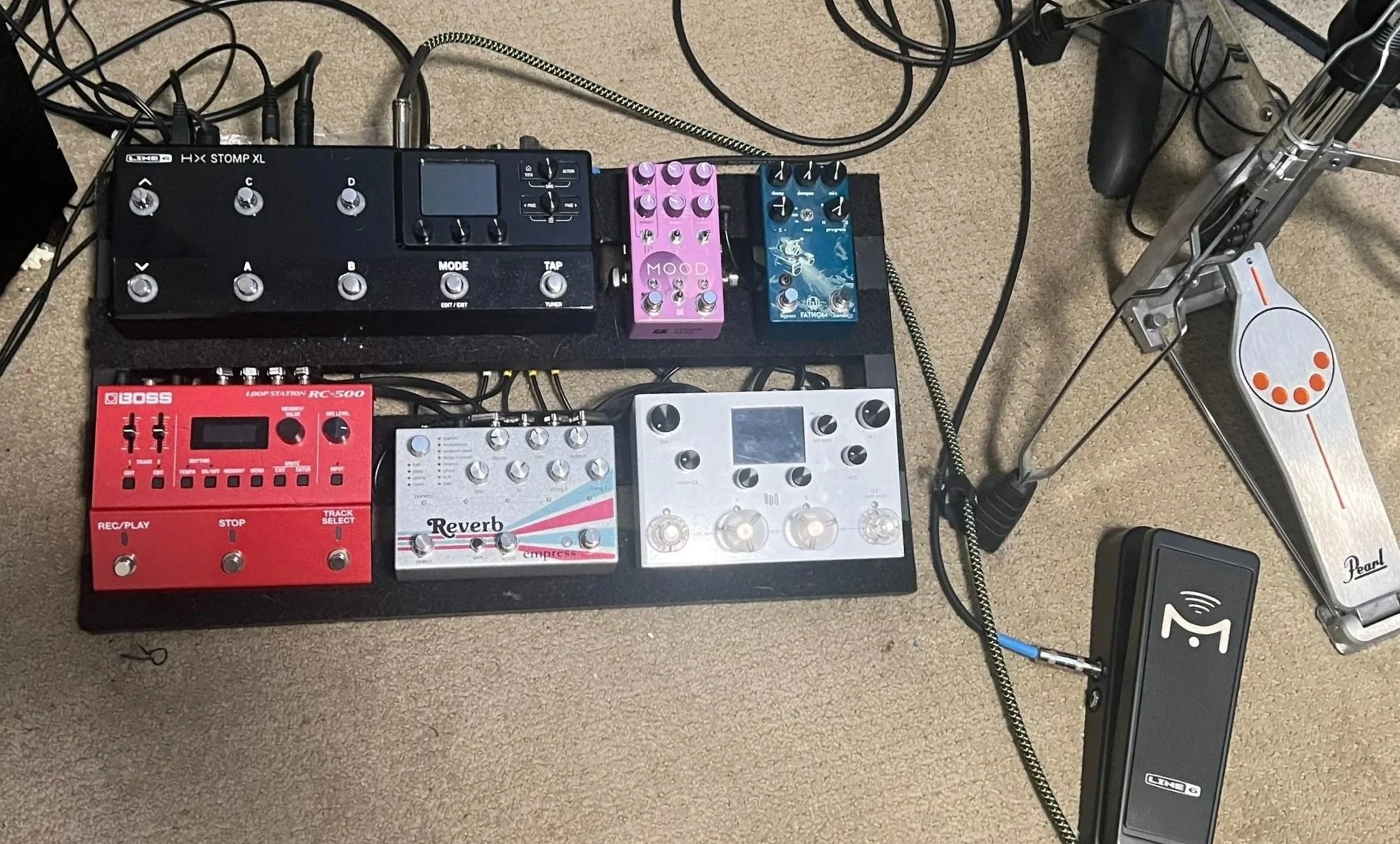 An overhead view of a guitar pedalboard with multiple effects pedals and a drum trigger pedal. The pedalboard includes a black HX Stomp XL, a pink Mood pedal, a blue Fathom pedal, a red Boss RC-500 loop station, a white Reverb pedal, and a white expression pedal on a beige carpeted floor. Numerous cables connect the pedals.