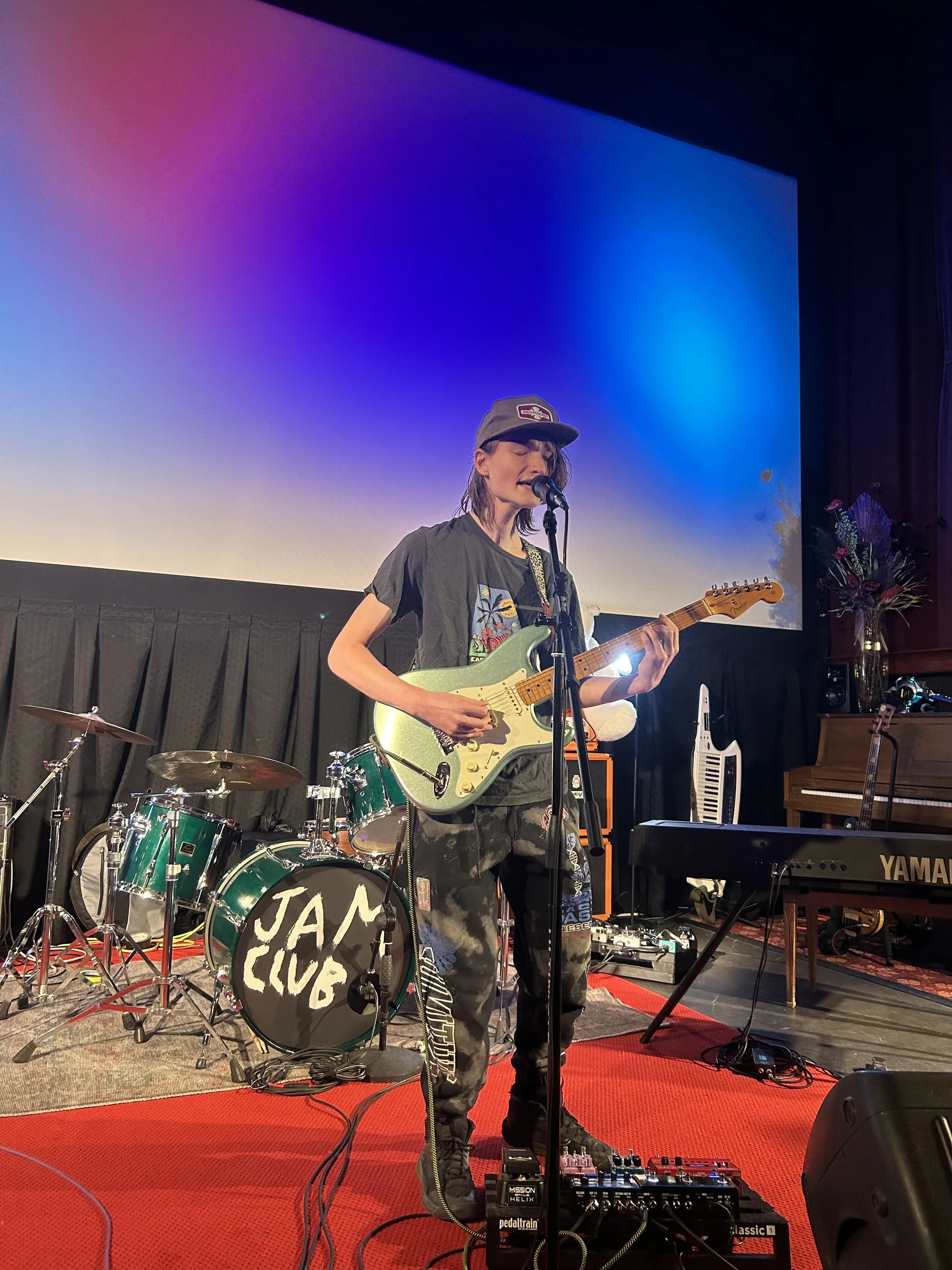 A young woman performs on stage with a microphone and electric guitar at a venue labeled 'JAM CLUB.' She is wearing a grey cap, a dark graphic t-shirt, and black cargo pants with patches. Behind her are drums, a keyboard, and some amplifiers, with a colorful backdrop on a large screen.