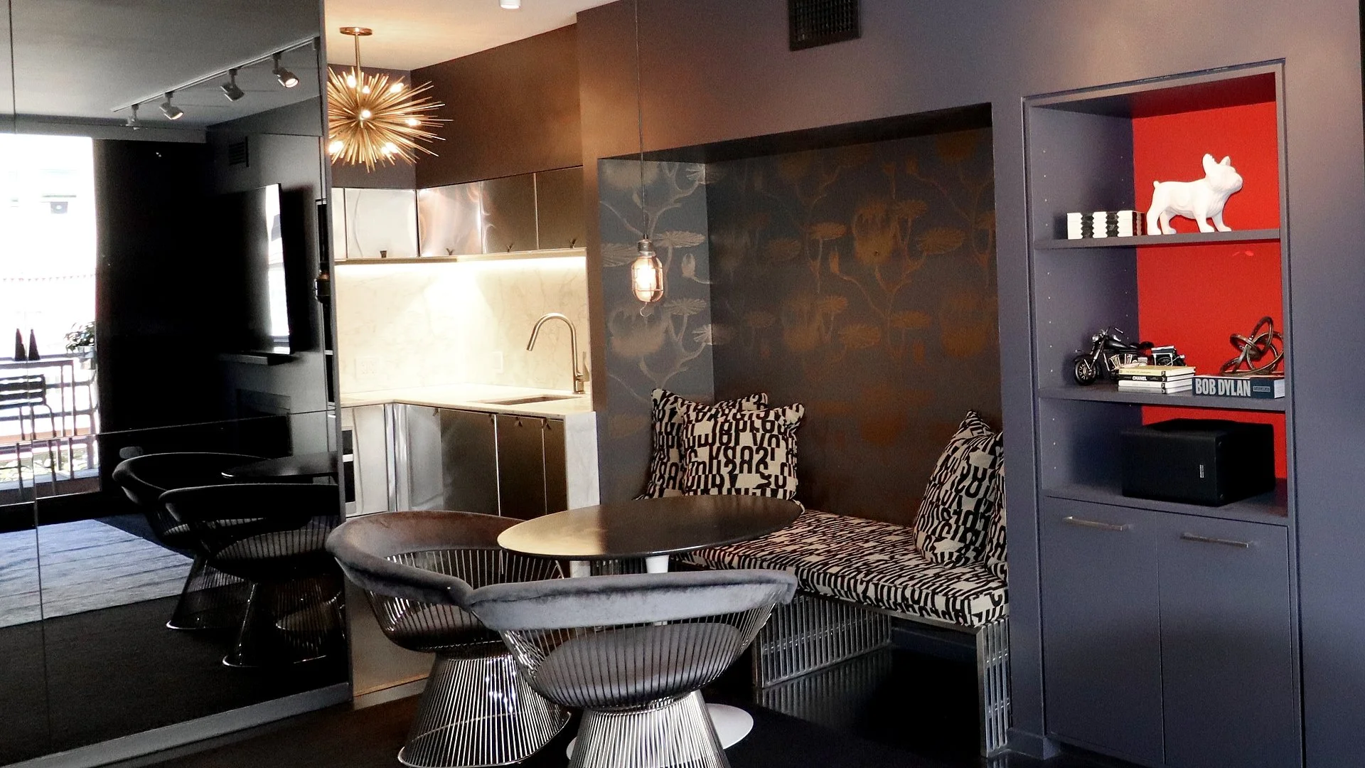 Modern living room area with black wire chairs, a black round table, a patterned bench with cushions, a grey built-in shelf with decorative items including a white bulldog statue, miniature motorcycle, books, and a speaker. A wall decorated with dark