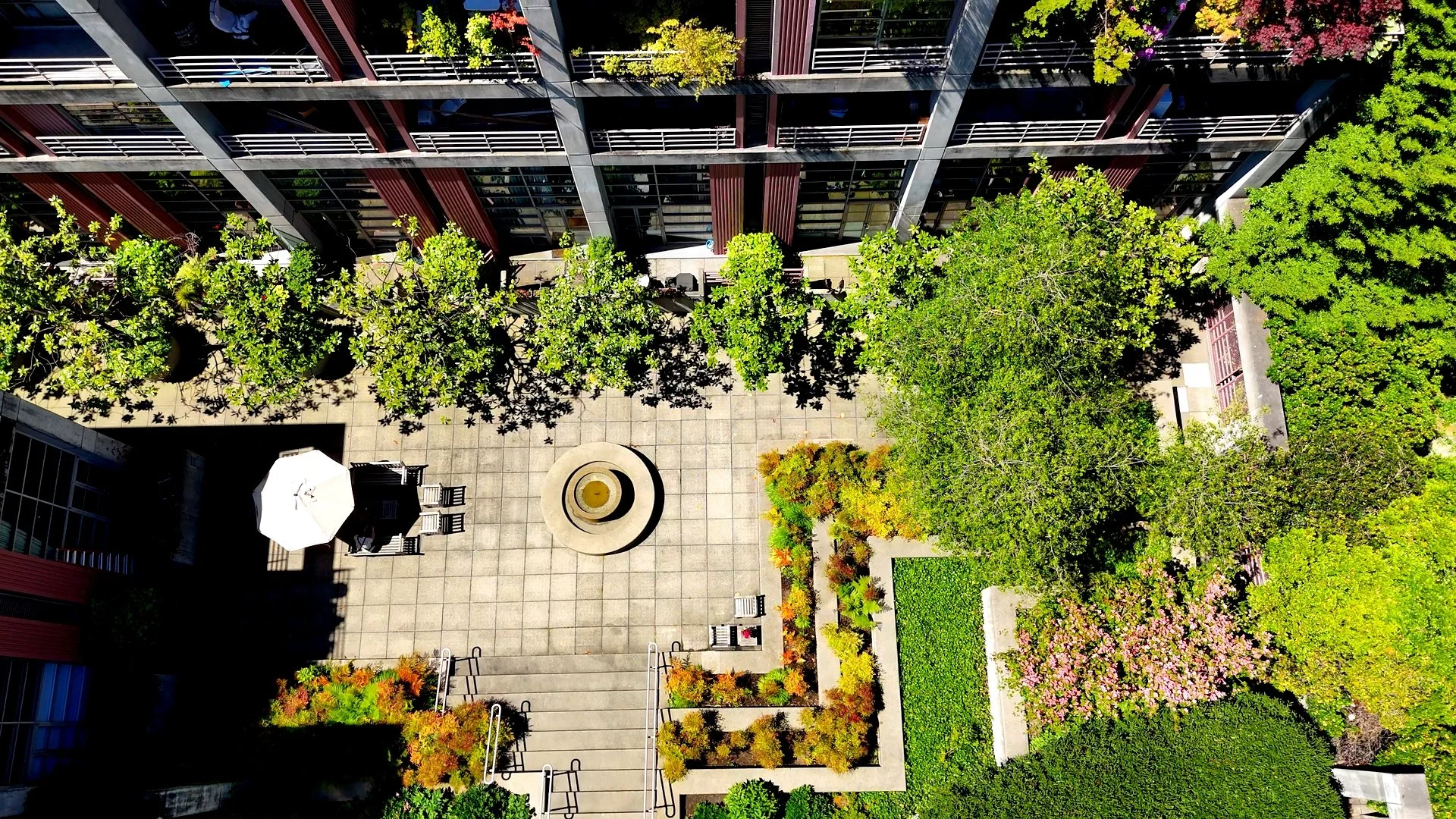 An aerial view of a city courtyard with planters, trees, a fountain, and an umbrella-shaded seating area surrounded by tall buildings.