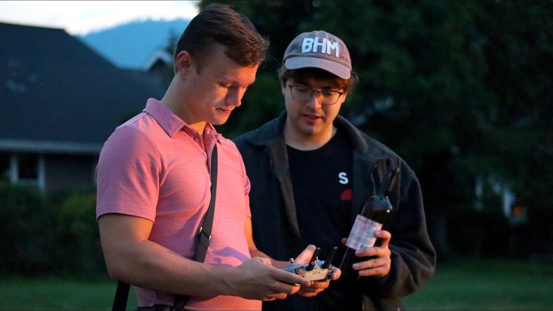 Two young men outdoors at dusk, one in a pink shirt and the other in a black jacket with a cap, are looking at a drone control remote and a bottle, with a house and trees in the background.