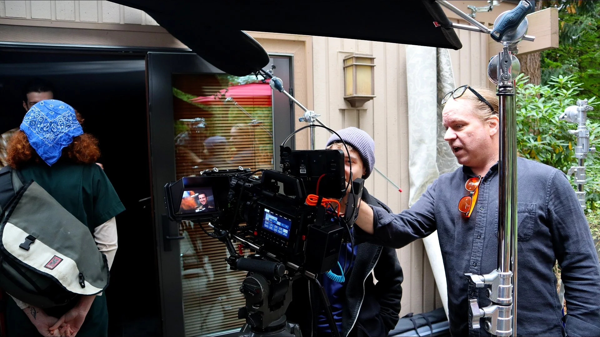 Film crew filming outdoors near wooden building with greenery, featuring a man with sunglasses on shirt, a woman with glasses and grey beanie, and a person with a blue bandana.
