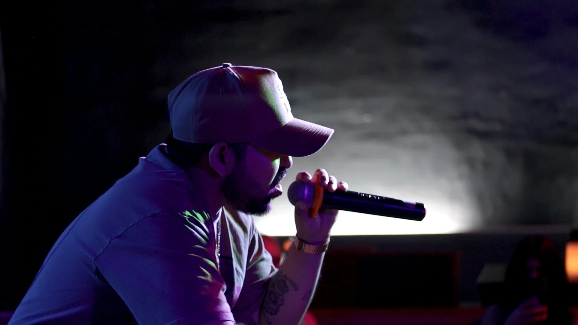 Man wearing a baseball cap and glasses, holding a microphone, appears to be singing or speaking, with dark background and dim lighting.