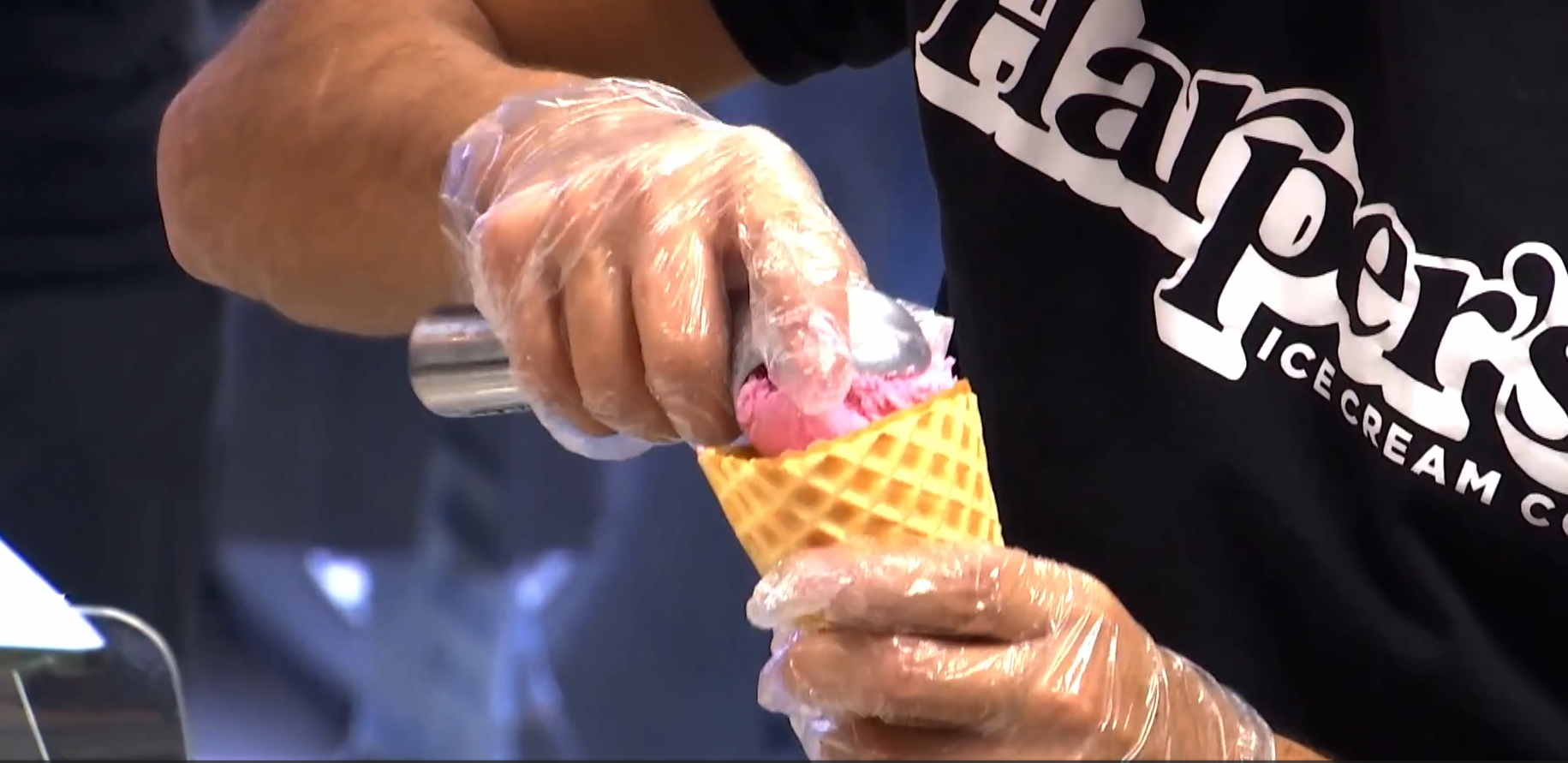Person making ice cream in a waffle cone, wearing plastic gloves, at an ice cream shop.