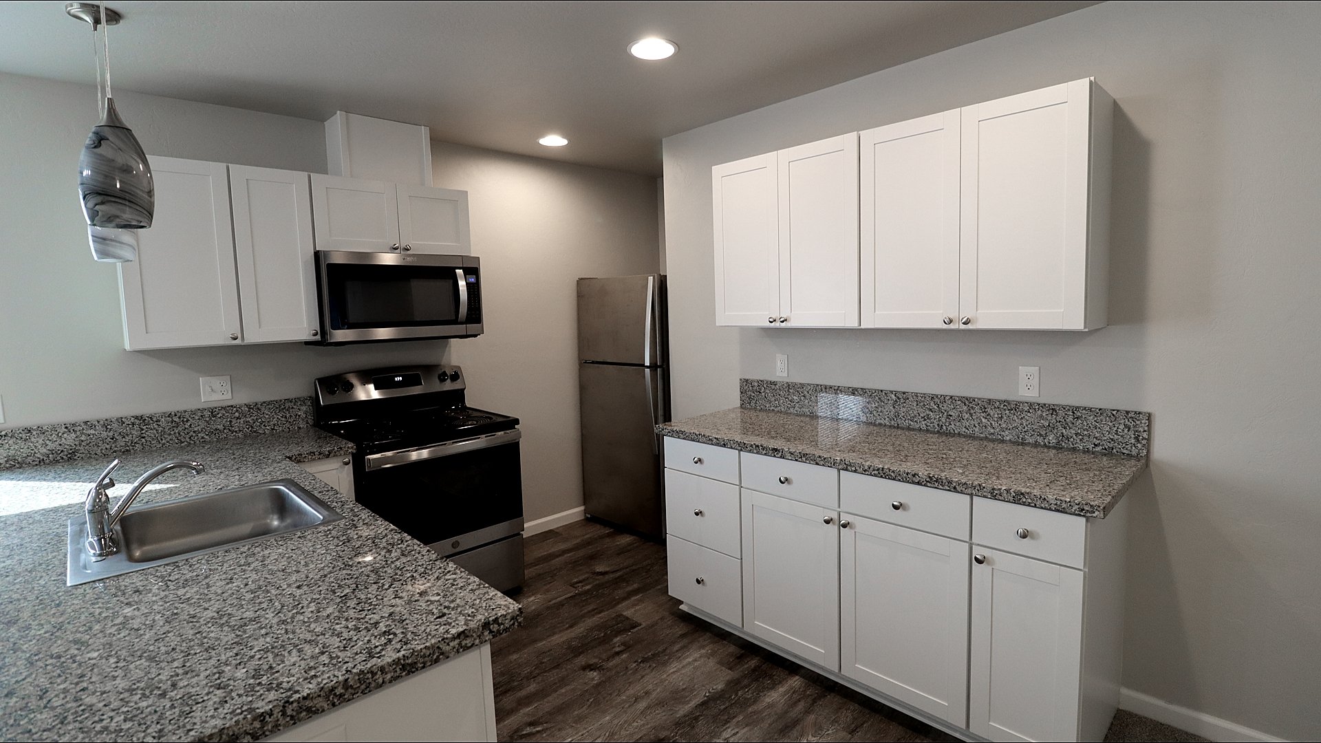 Modern kitchen with white cabinets, granite countertops, stainless steel appliances including a microwave, stove, and refrigerator, a sink with a chrome faucet, and wood-look flooring.
