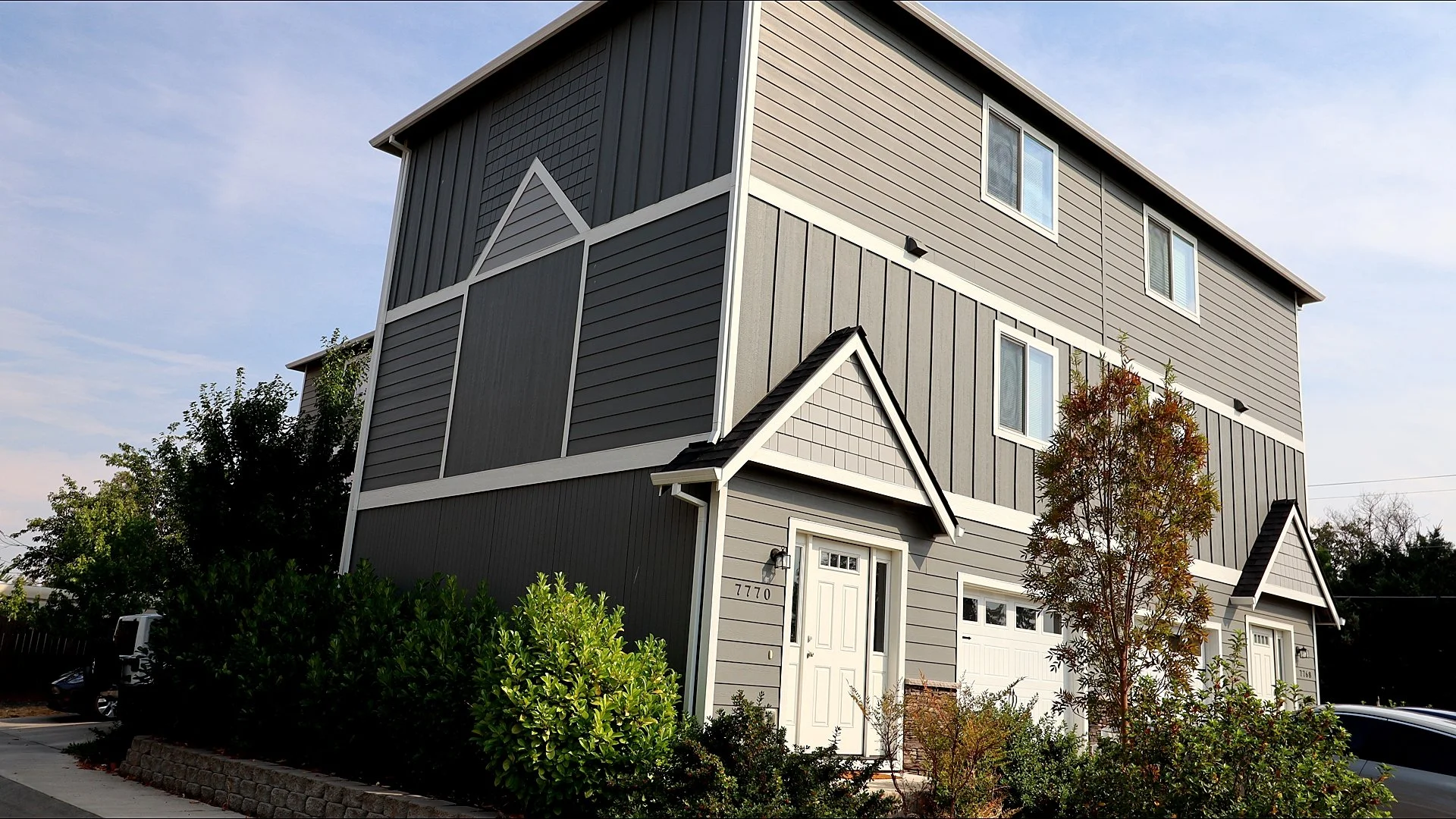 A modern, multi-story house with gray siding, white trim, and multiple windows, surrounded by greenery and small trees.