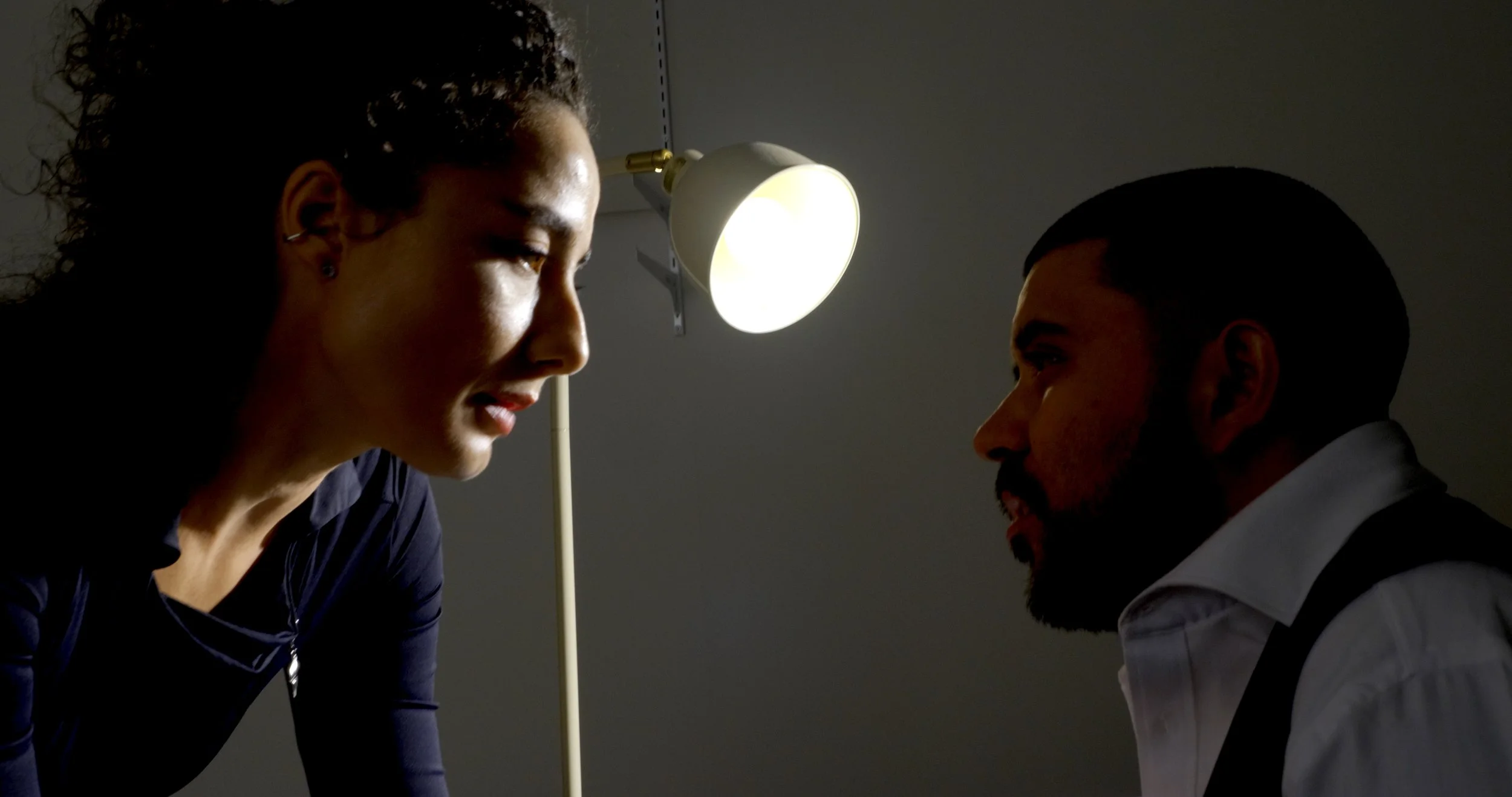 A woman leaning forward face-to-face with a man in a dimly lit room next to a bright standing lamp.