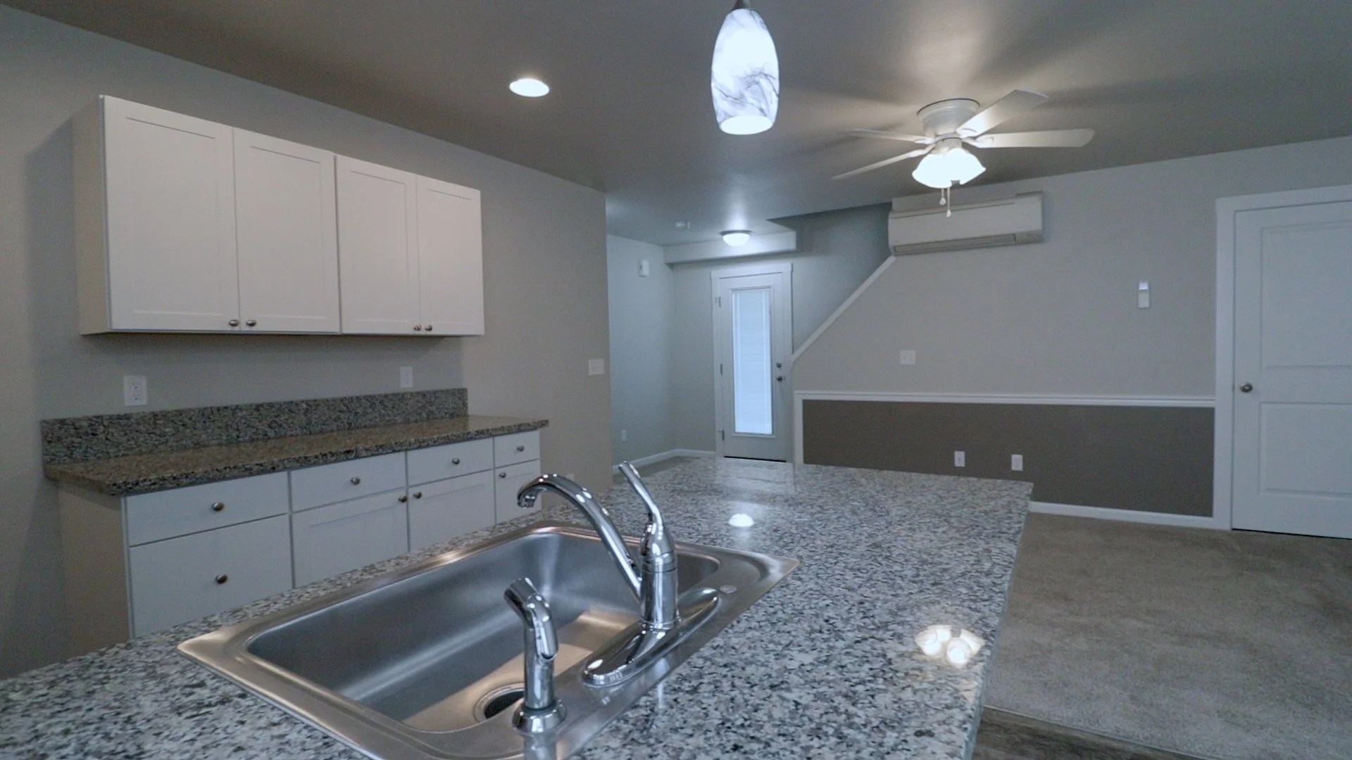 Interior view of a modern kitchen with granite countertops, white cabinets, stainless steel sink, and a ceiling fan. A door leads outside, and there's an air conditioning unit on the wall.