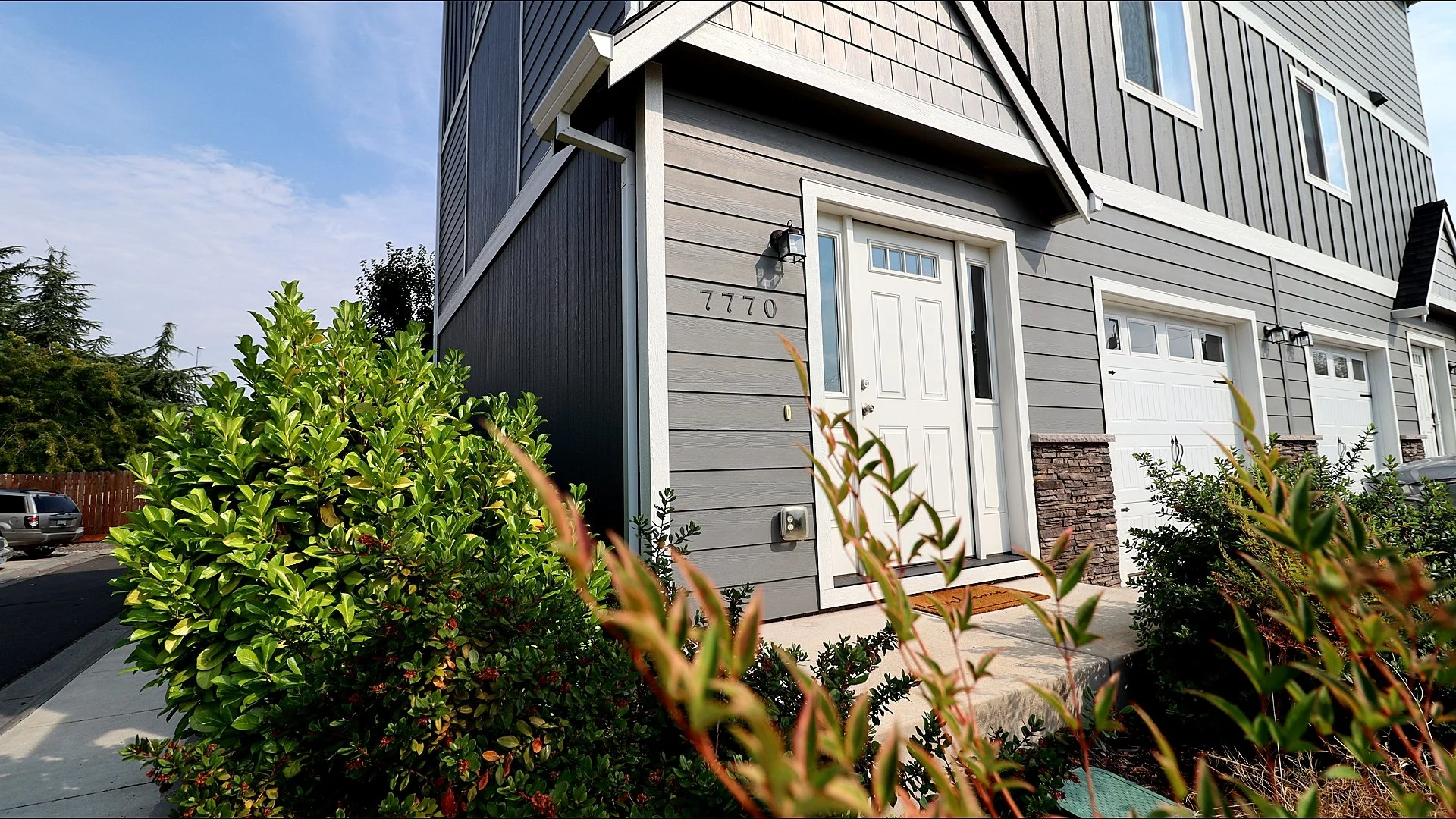 Front view of a modern townhouse with gray siding, white door, and garage, with bushes and plants in the foreground, and a parking lot with cars to the left.