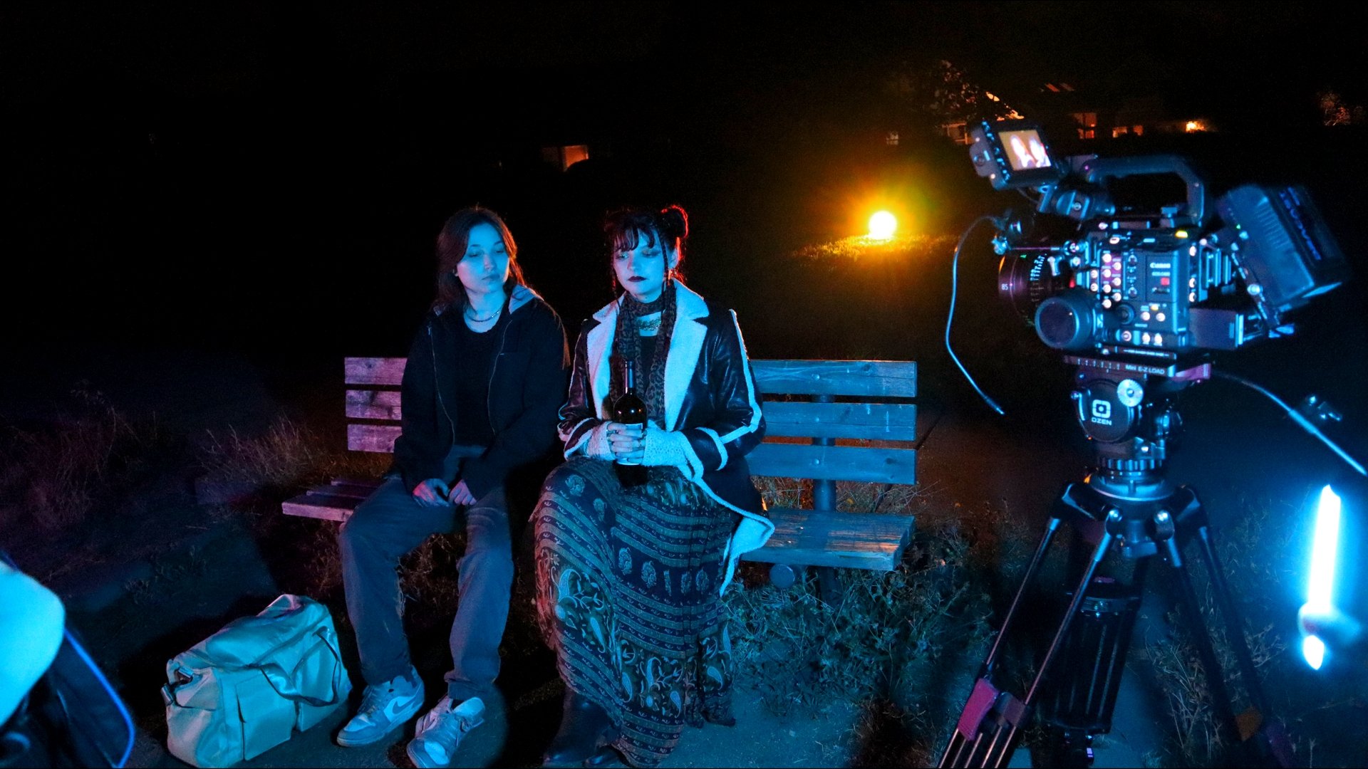 Two young women sitting on a bench at night in front of a camera on a tripod recording or photographing them. The woman on the left has dark hair and is wearing a black hoodie and gray pants. The woman on the right has dark hair in pigtails, is weari