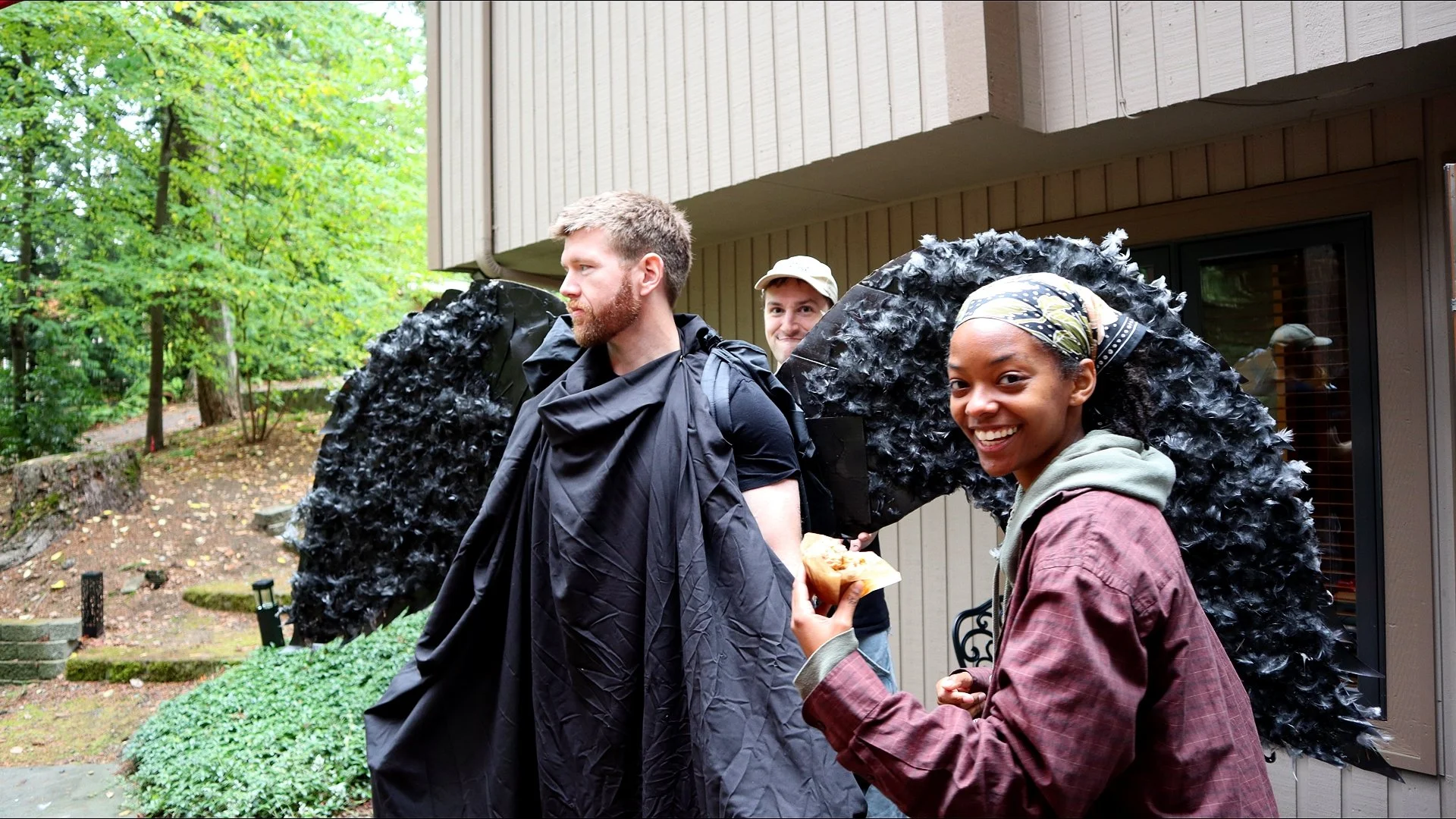 Three people, two women and one man, dressed as angels with large black wings, standing outside near a house with trees in the background. The woman in the foreground is smiling, holding food, and wearing a headscarf. The man is looking to the side, 