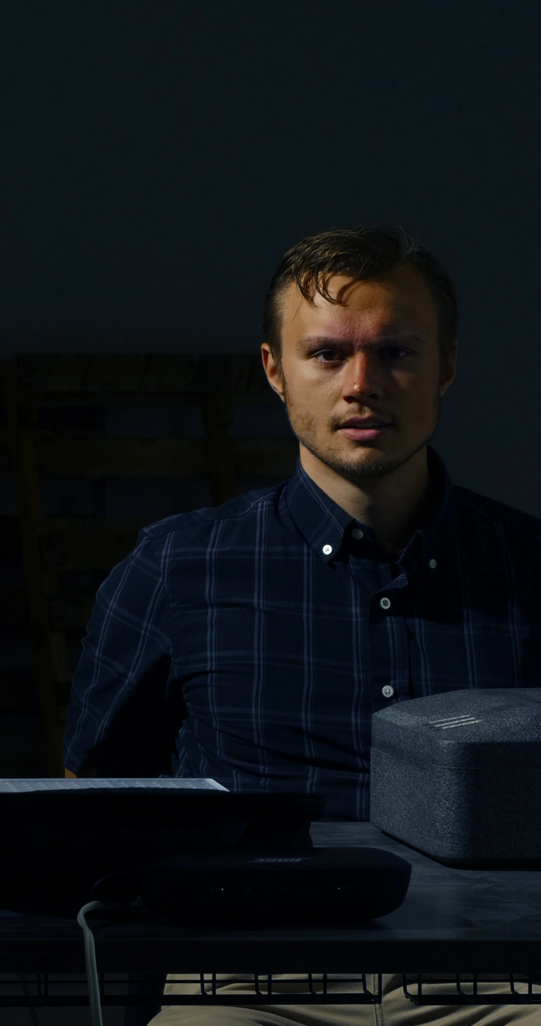 A man with light skin and brown hair, wearing a blue plaid shirt, sitting at a desk with a speaker and some other electronic devices, in a dimly lit room.