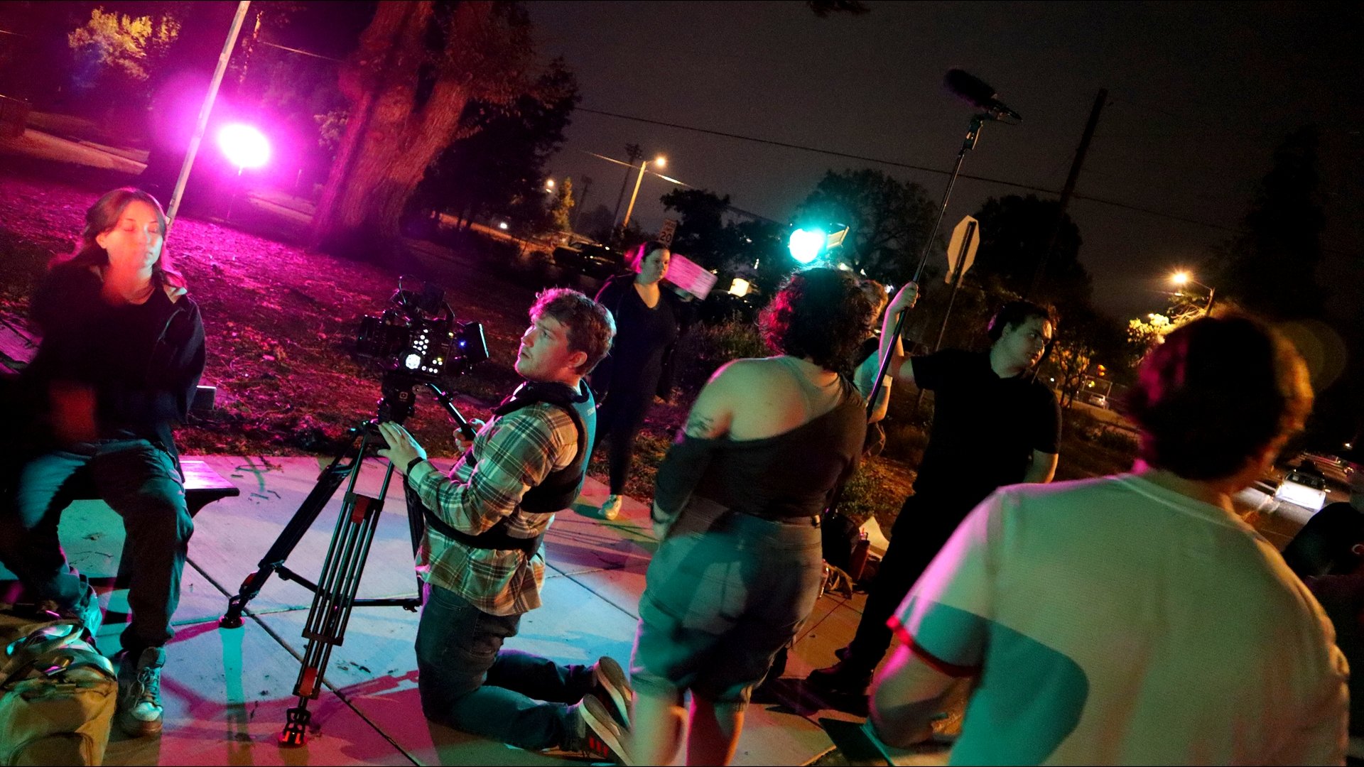 A film crew setting up equipment outdoors at night, with colorful lighting, producing a scene involving a woman sitting on a bench and several crew members behind the scenes.