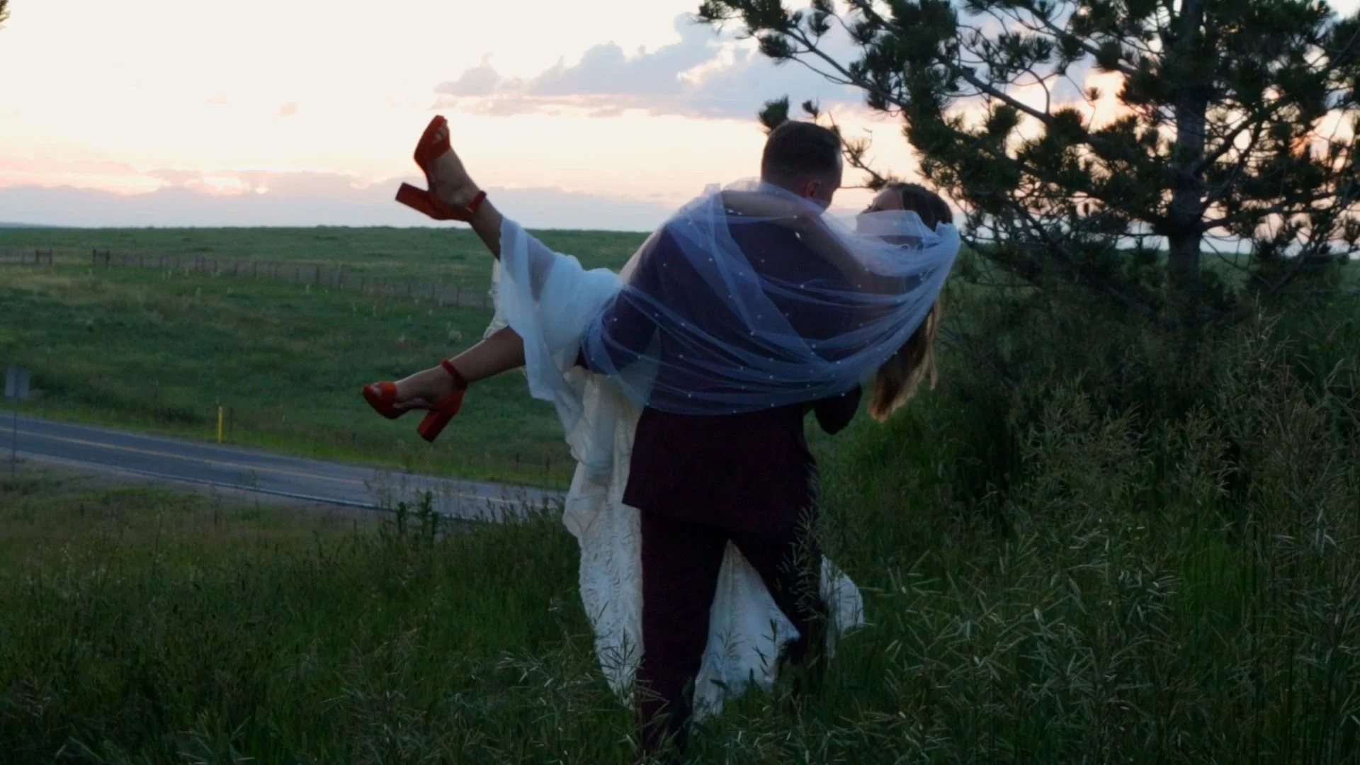 A groom carrying a bride dressed in a wedding gown and high heels through a grassy field at sunset.