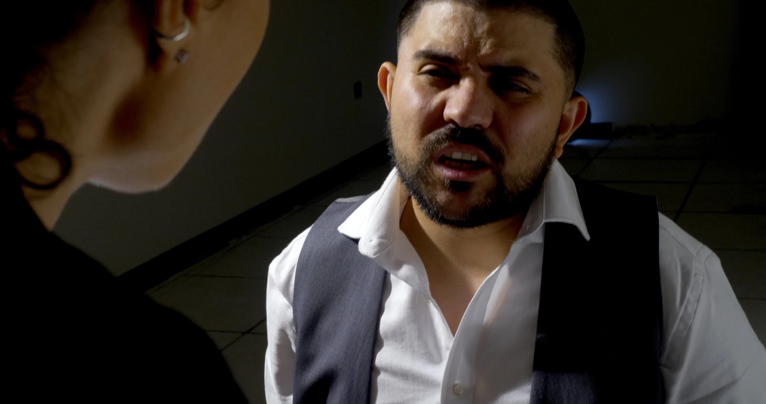A close-up of a man with a beard and short dark hair, wearing a white shirt and a vest, appearing to be angry or upset during a confrontation with a woman whose face is partly visible with earrings and tattoos on her neck.