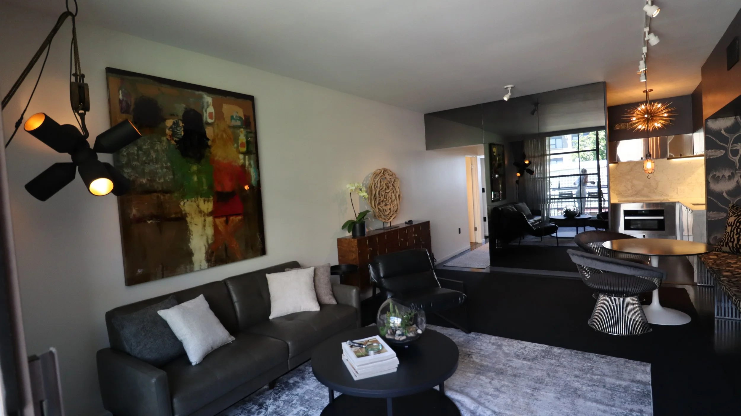 Modern living room with a black leather sofa, a large colorful abstract painting on the wall, a black ceiling fan light, a round coffee table with magazines and a glass terrarium, a dark wood sideboard with a white orchid, and a dining area with a ro