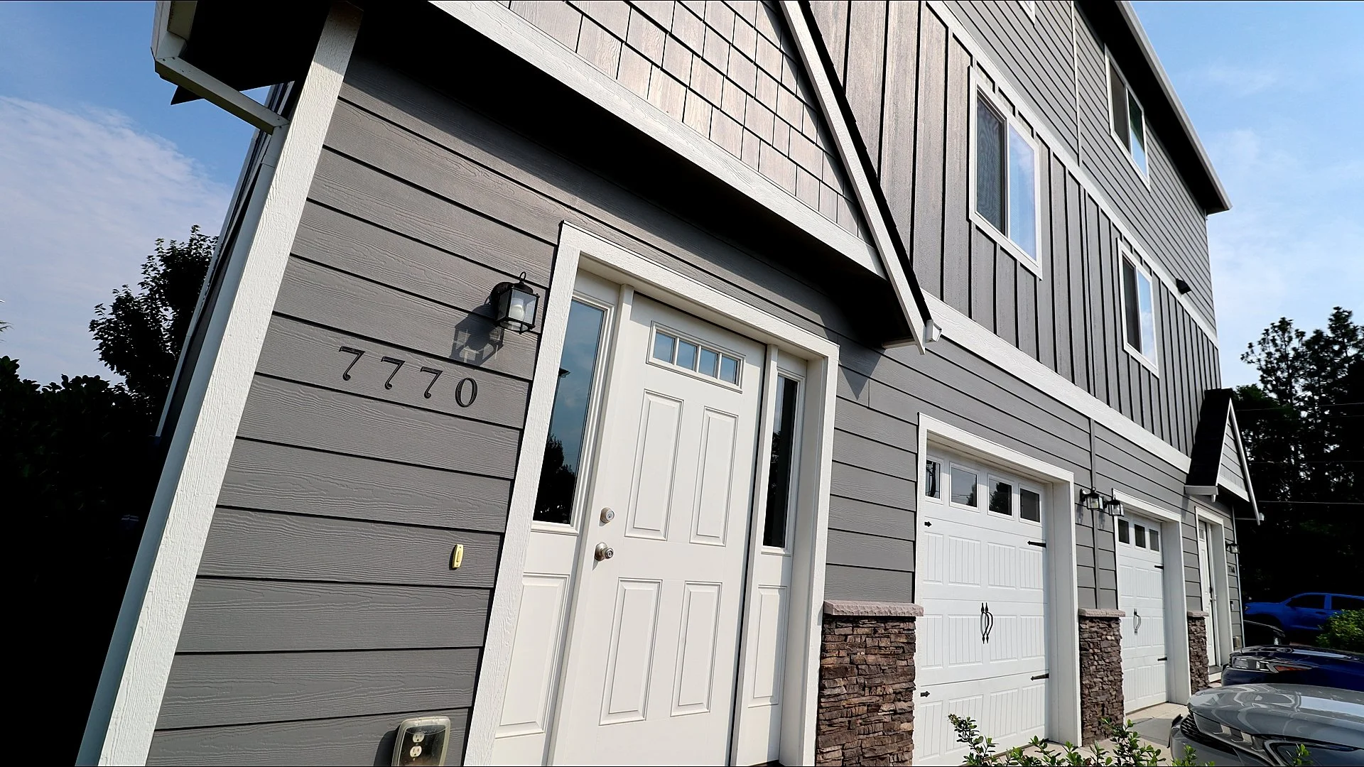 Front view of a modern residential building with gray horizontal siding, white doors, and garage doors, and house number 7770 on the wall.