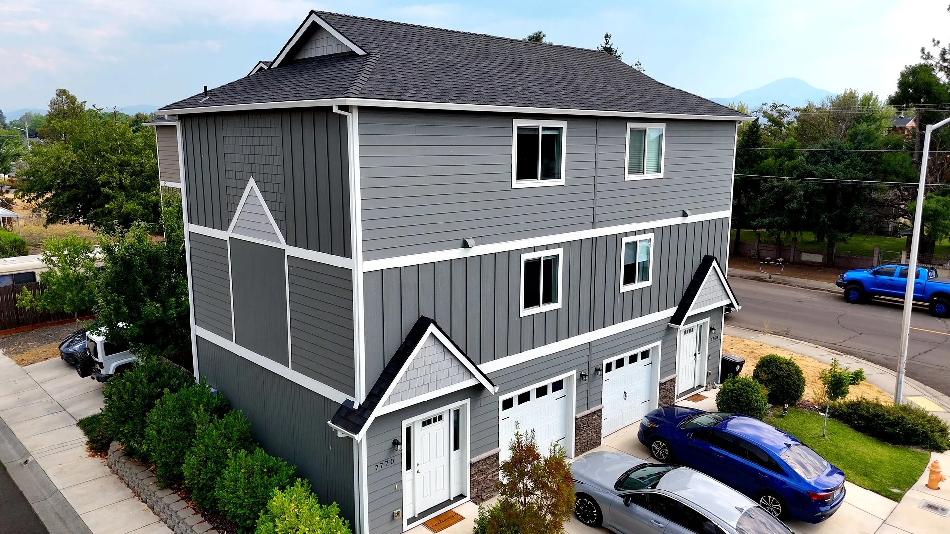 A modern, three-story gray townhouse with white trim, attached garage, and surrounding cars and landscaping.