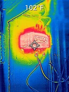 Infrared thermal image of an electrical panel with temperature reading of 102°F, showing hot spots in red and pink, cooler areas in blue and green.