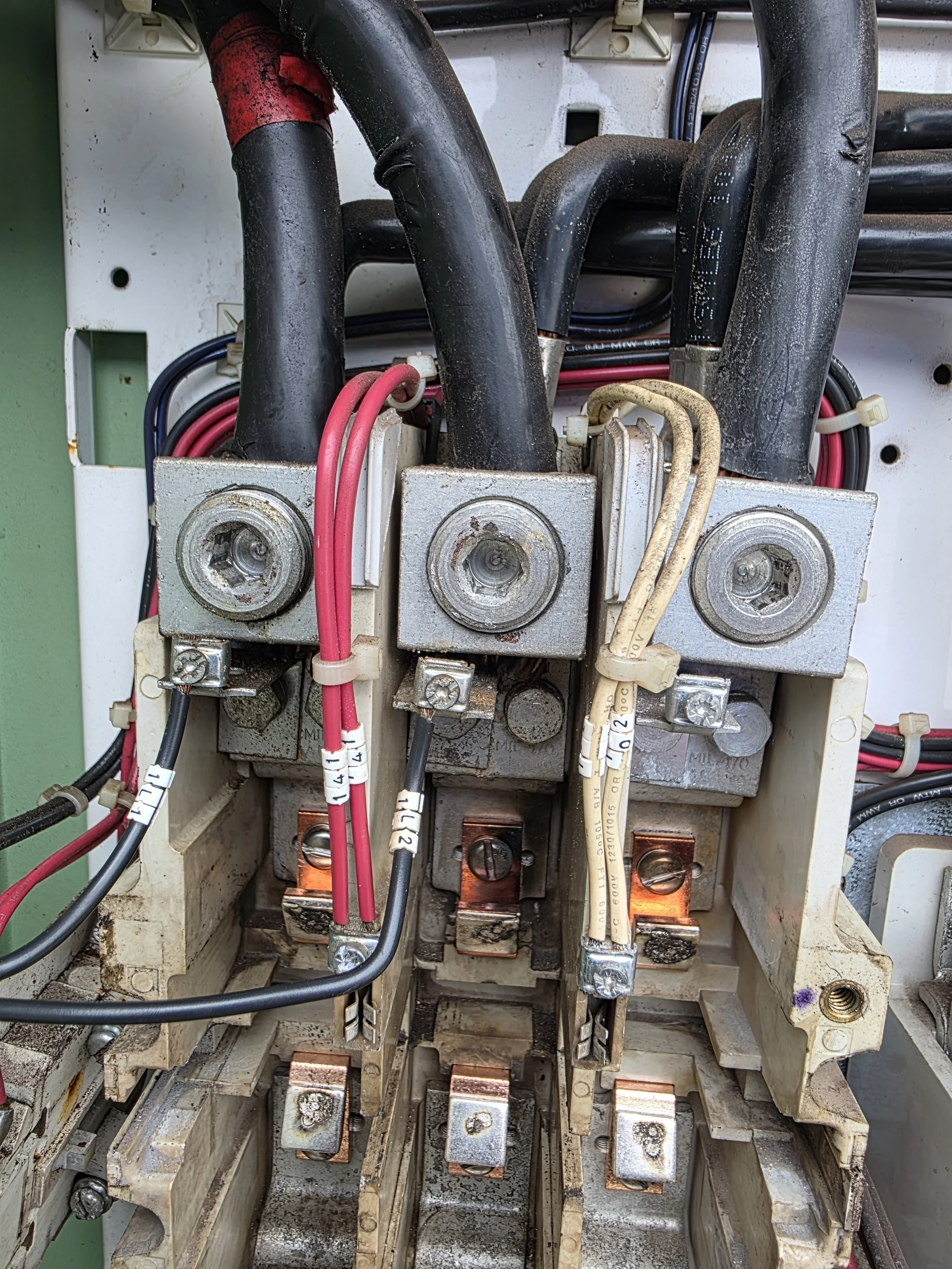 Close-up view of internal electrical components, including three large electrical contactors with electrical wires connected, inside an electrical panel or control box. The components are mounted on a dirty metal frame with visible screws.