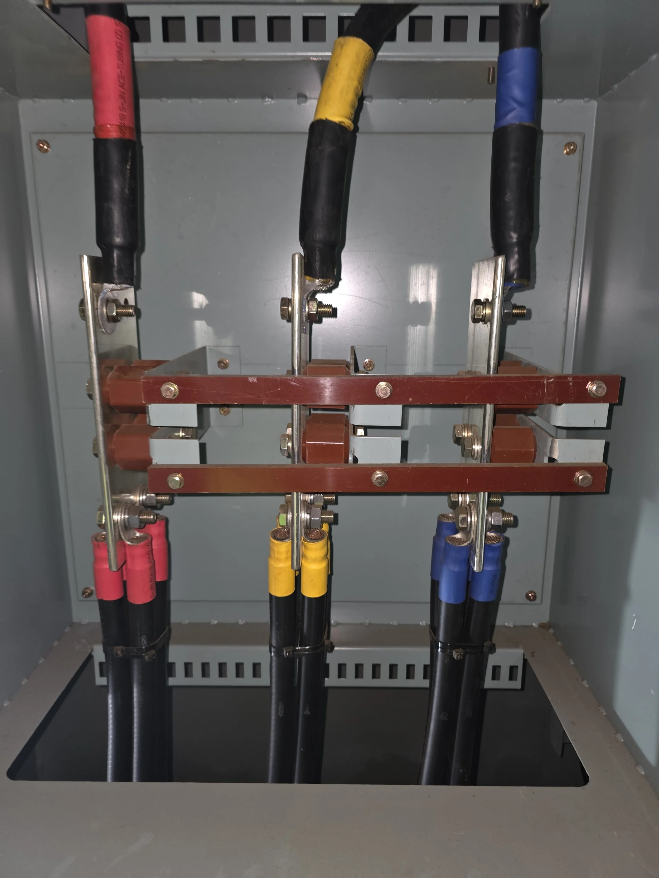 Inside an electrical panel with three large insulated cables connected to bus bars, each cable covered with red, yellow, and blue insulation. The panel has a metal interior with mounting screws and a vented cover at the top.