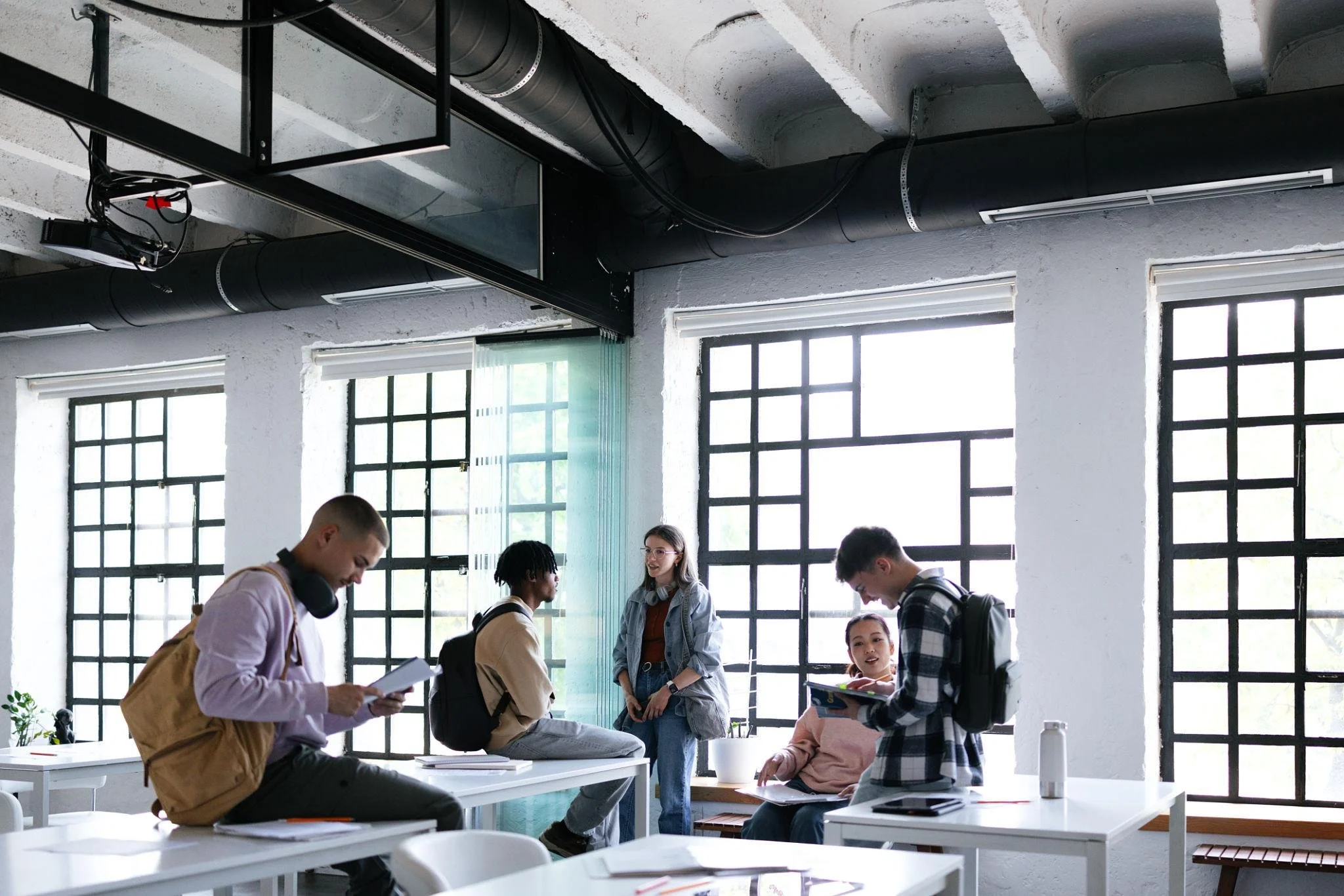 Five students in a modern classroom with large windows and geometric black window frames, engaging in conversation and studying.