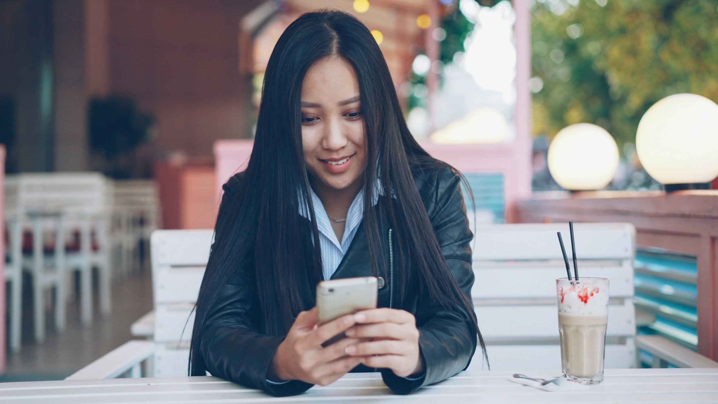 A young woman with long dark hair, wearing a black leather jacket and a blue shirt, sitting at an outdoor cafe table, looking at her smartphone with a smile. There is a milkshake with strawberries and two straws on the table.