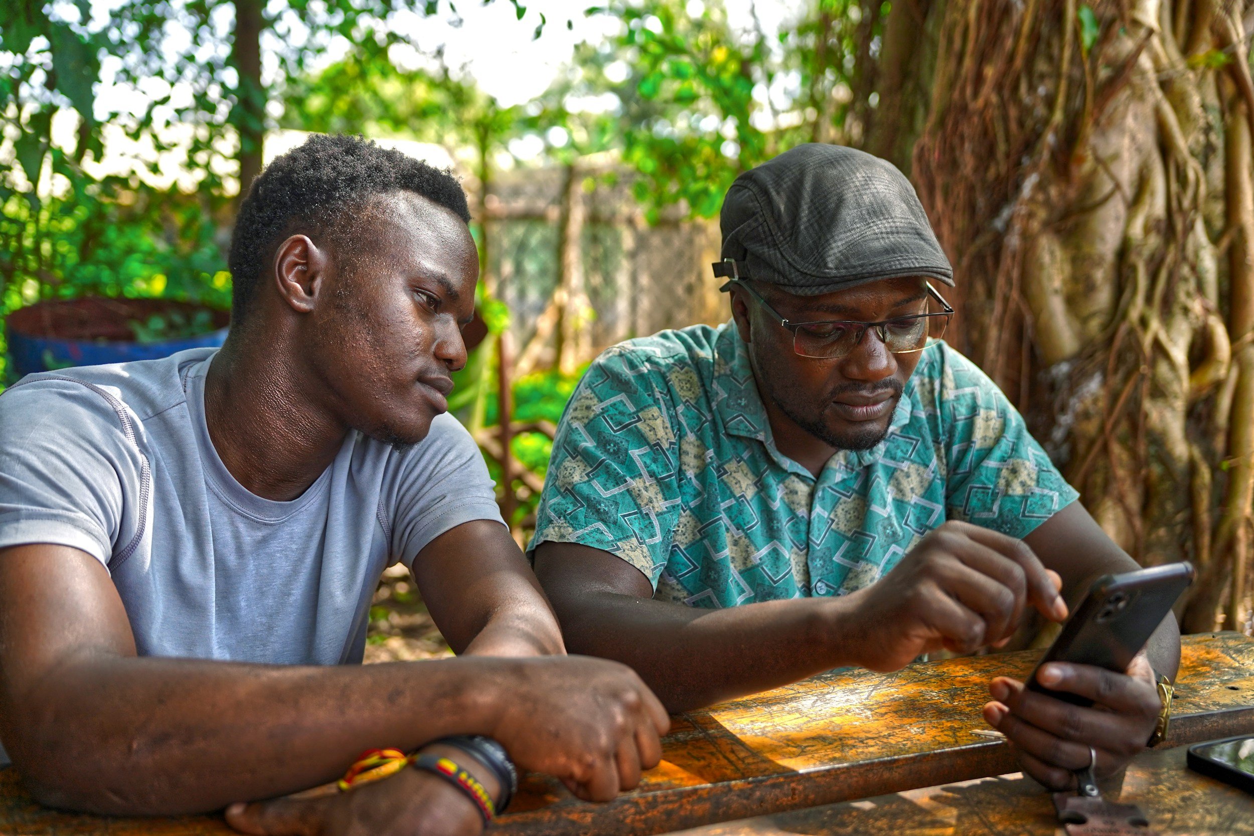 Two men sitting at a wooden table outdoors, one is looking at a smartphone while the other looks on, with a lush green background and large tree trunk.