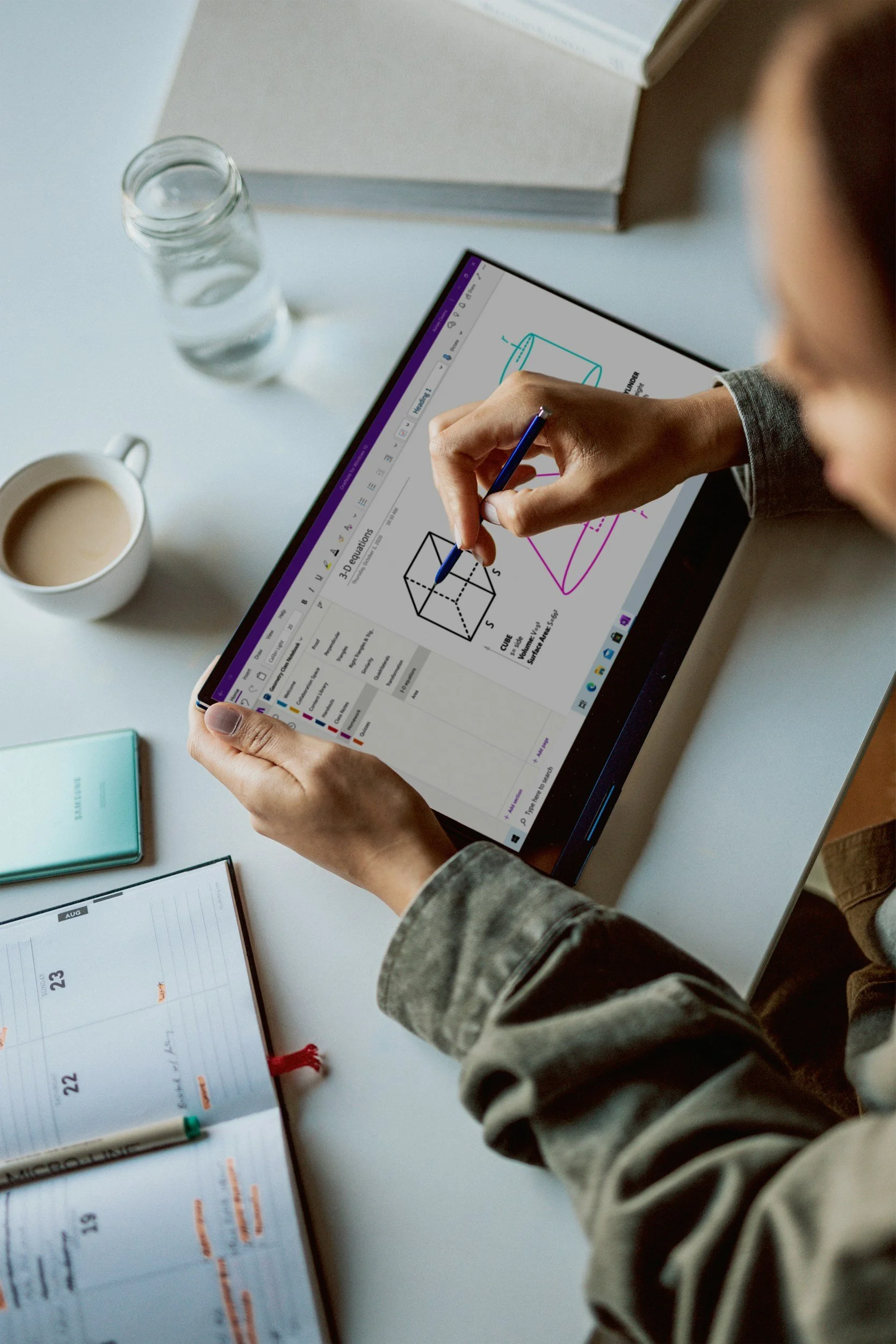 A person working on a digital tablet displaying 3D geometric diagrams, with a cup of coffee, a glass of water, a planner, and a notebook on the desk.