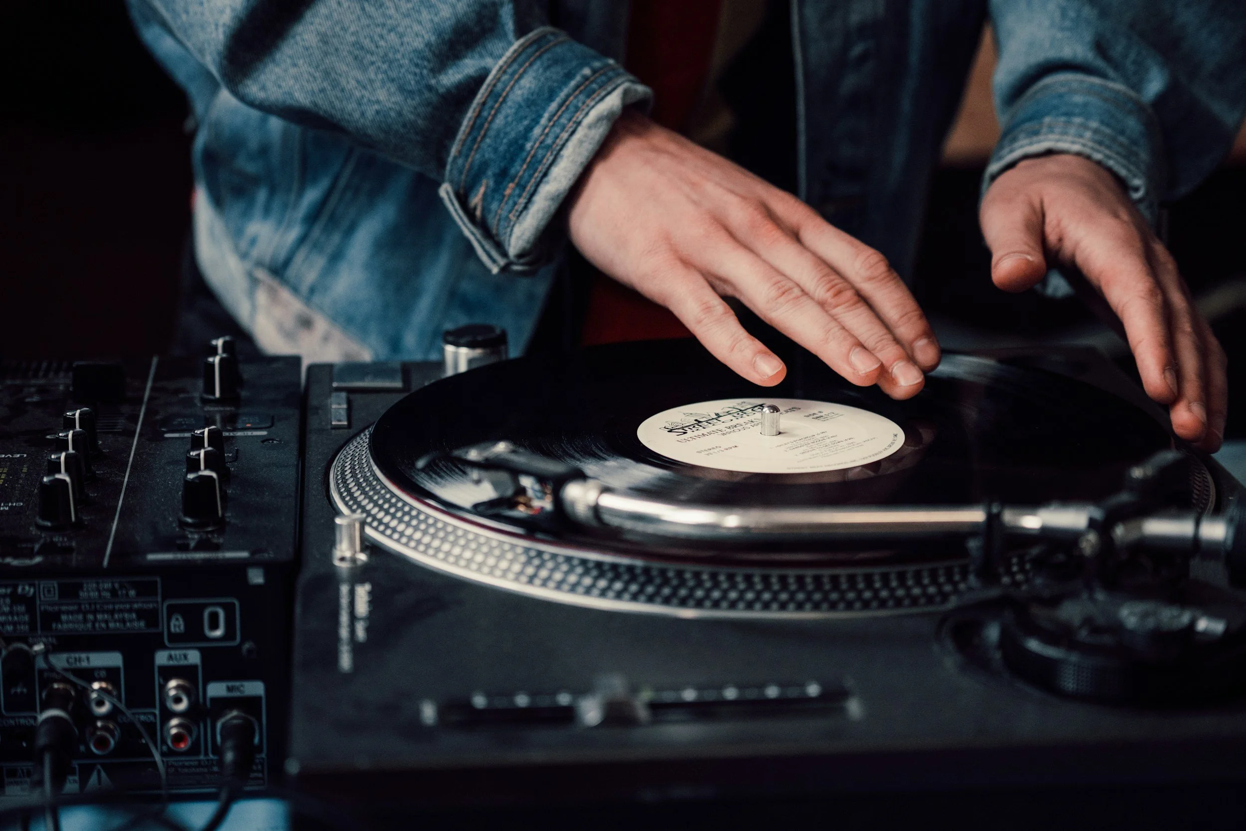 Person in a denim jacket using a turntable to DJ, with visible controls and vinyl record.