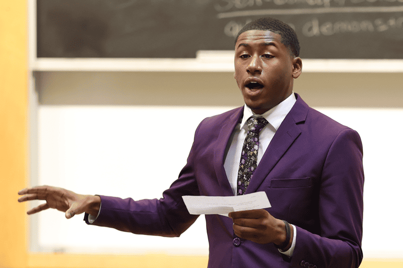 Young man in purple suit giving a presentation in a classroom.