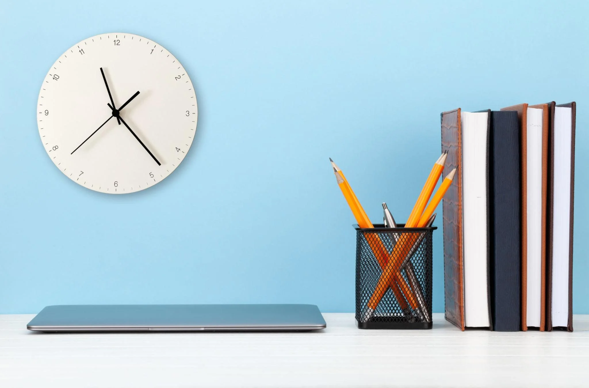 A minimalist workspace with a closed silver laptop, a black mesh pen holder containing orange pencils and a silver pen, and a row of five vertically standing notebooks with different covers against a light blue wall with a white clock showing approximately 10:54.
