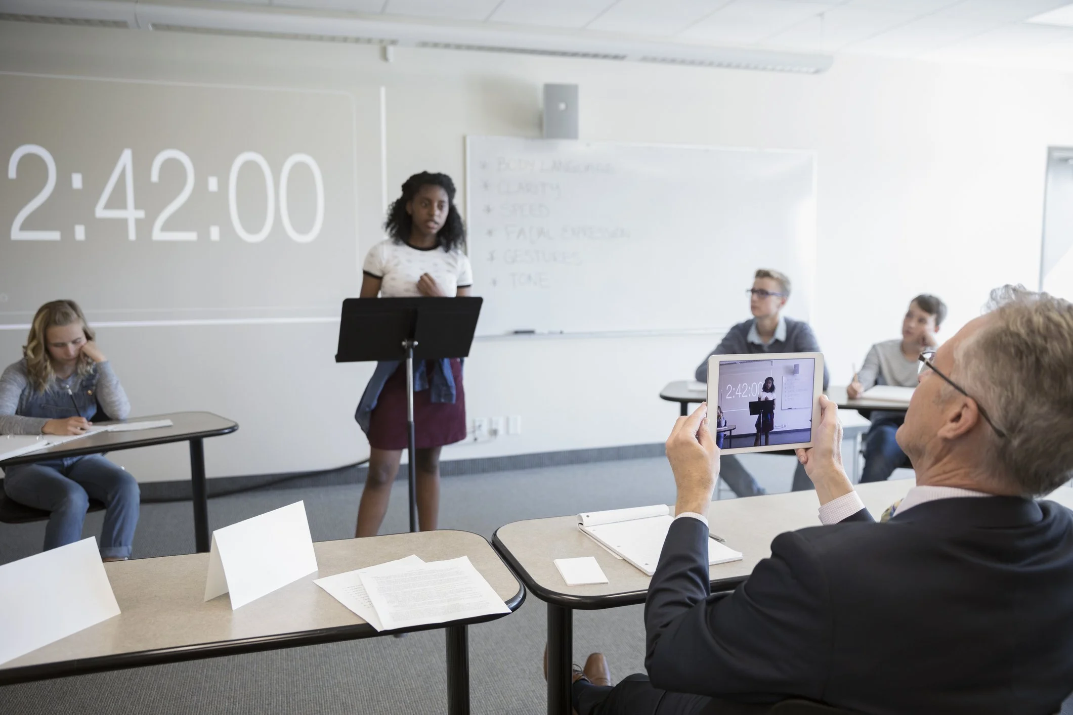 A classroom scene with a girl presenting at a podium, a timer on the wall showing 2:42:00, and a man recording the presentation on a tablet. Other students are seated at desks, listening and taking notes.