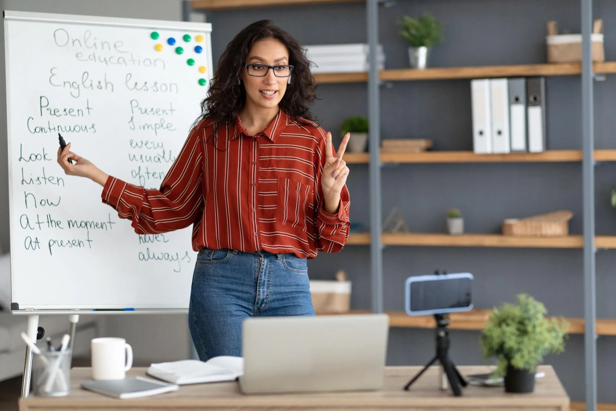 A woman teacher standing in front of a whiteboard with English lesson notes, gesturing with her right hand holding a pen and making a peace sign with her left hand, during an online lesson.
