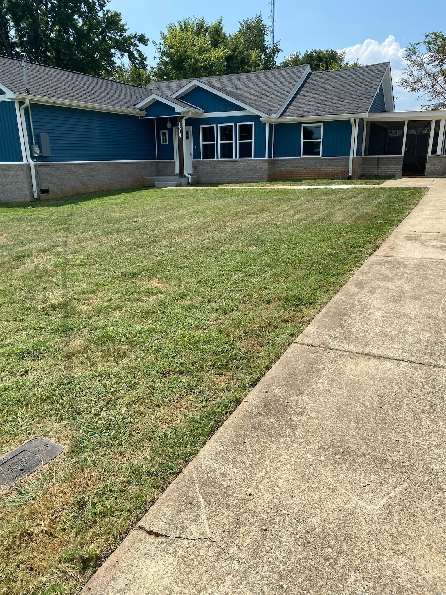 Front yard and exterior of a blue house with a concrete sidewalk and green grass, trees in the background, under a partly cloudy sky.