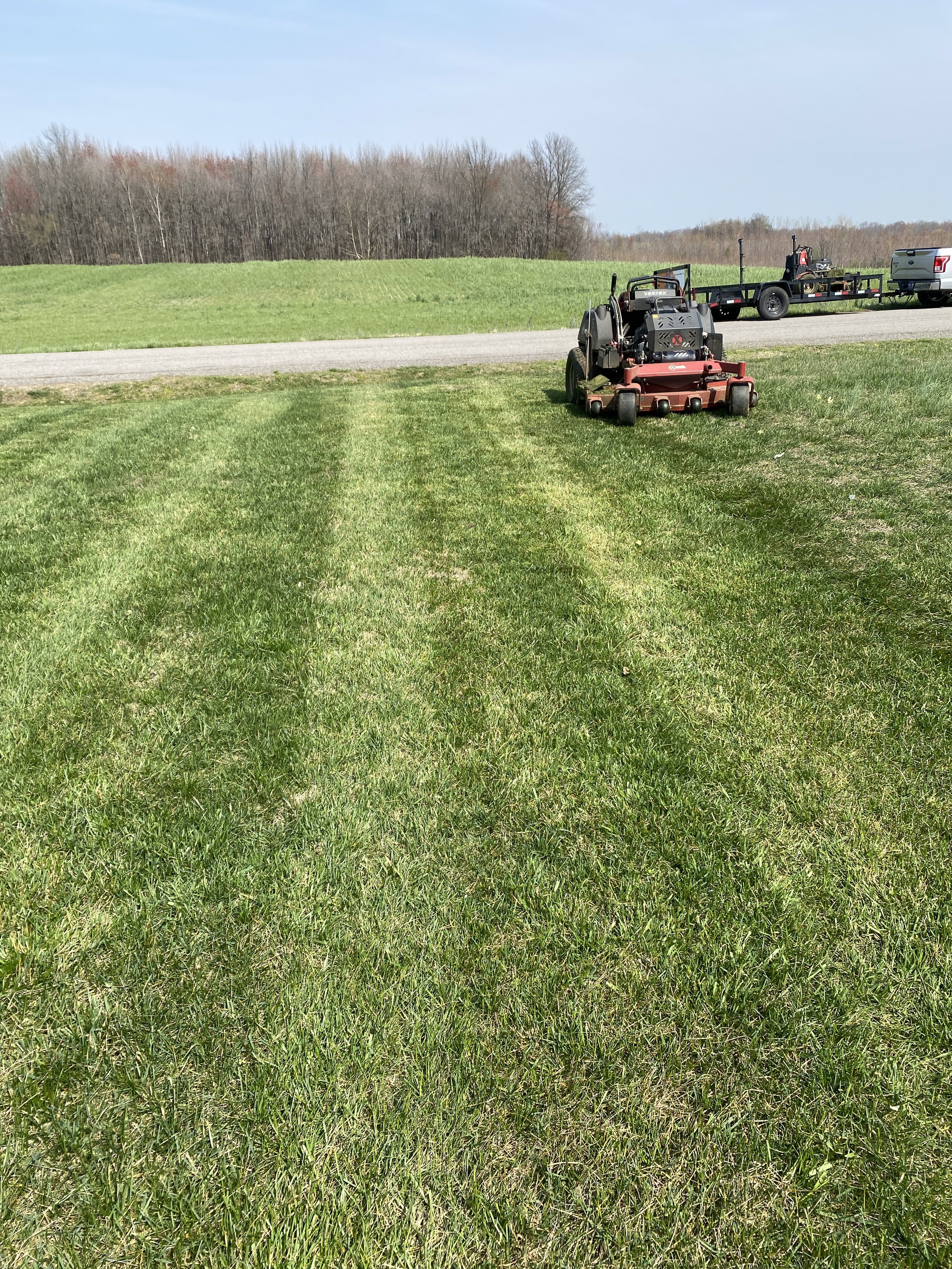 A lawn mower on a freshly mowed grassy area with a trailer and vehicle in the background, under a clear sky.