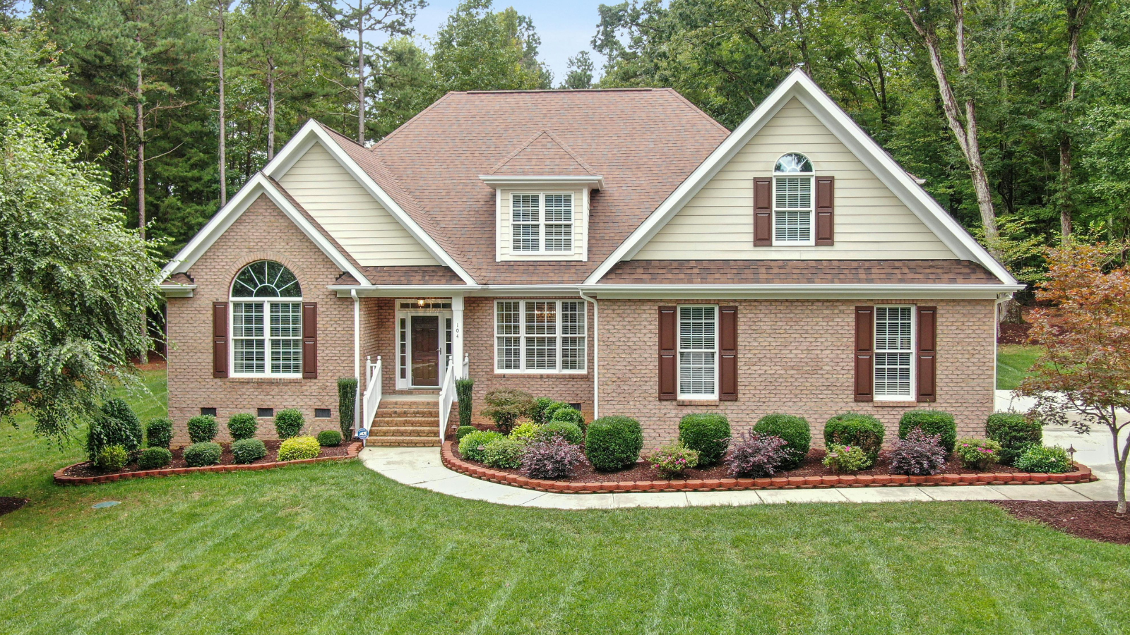 A two-story suburban house with a manicured front yard and garden, brick and siding exterior, gable roof, and multiple windows with brown shutters, surrounded by trees.