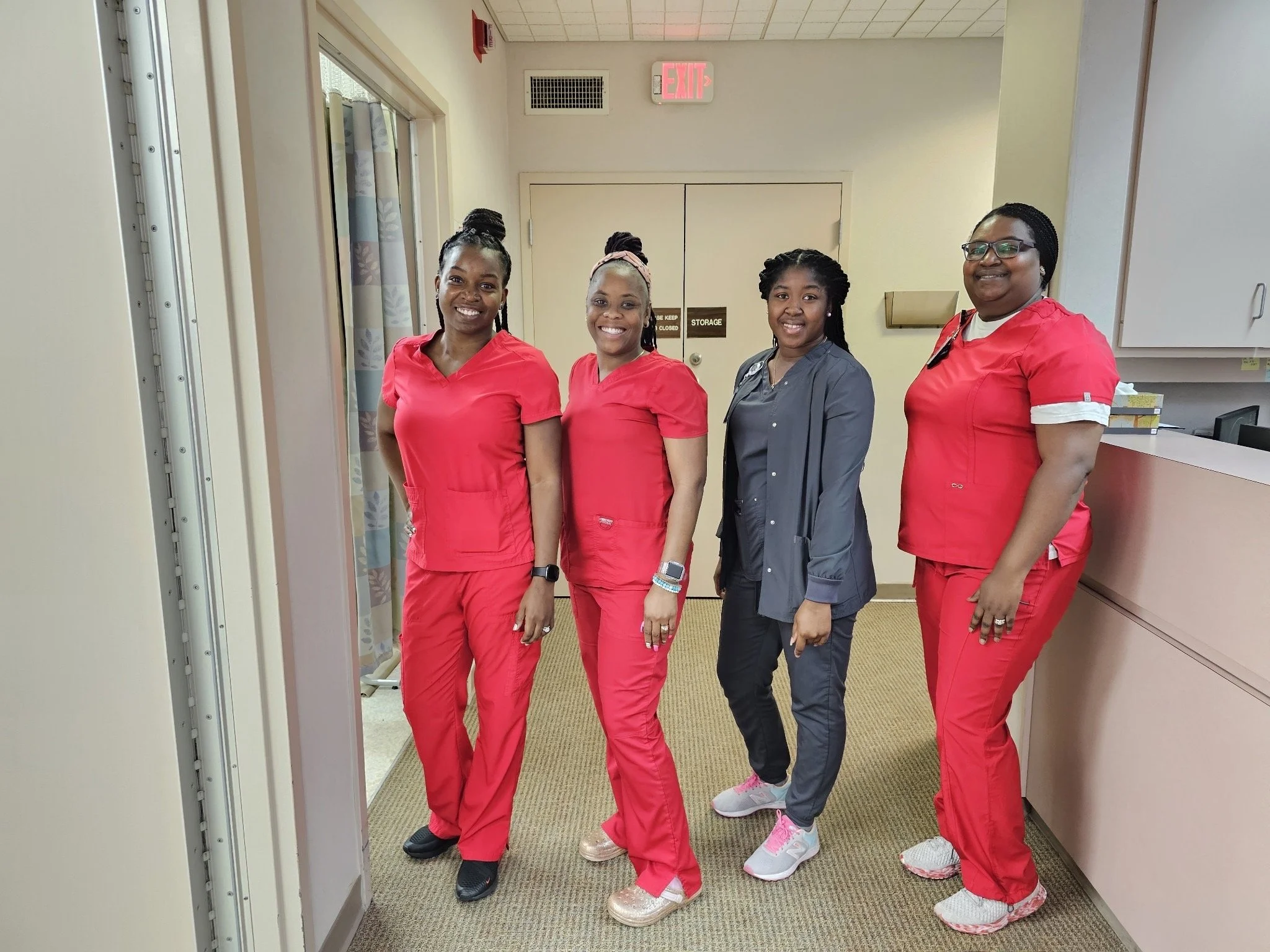 Four healthcare workers, three women in red scrubs and one woman in gray scrubs, standing together in a hospital corridor, smiling at the camera.