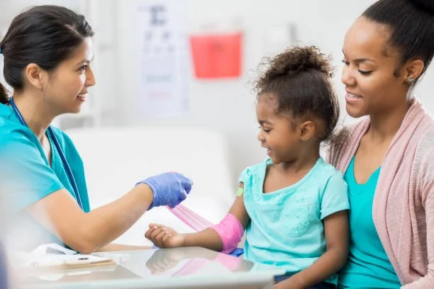 A healthcare professional in scrubs and gloves showing a pink bandage to a young girl with an Arm injury, sitting with her mother in a medical office.