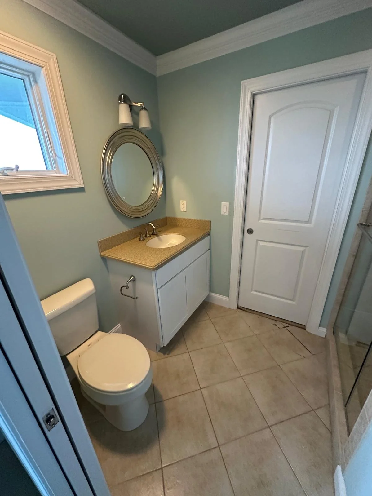 A small bathroom with a toilet, a vanity with a sink, a mirror above the sink, a small window, and a shower stall on the right. The walls are painted light green and the floor is tiled.
