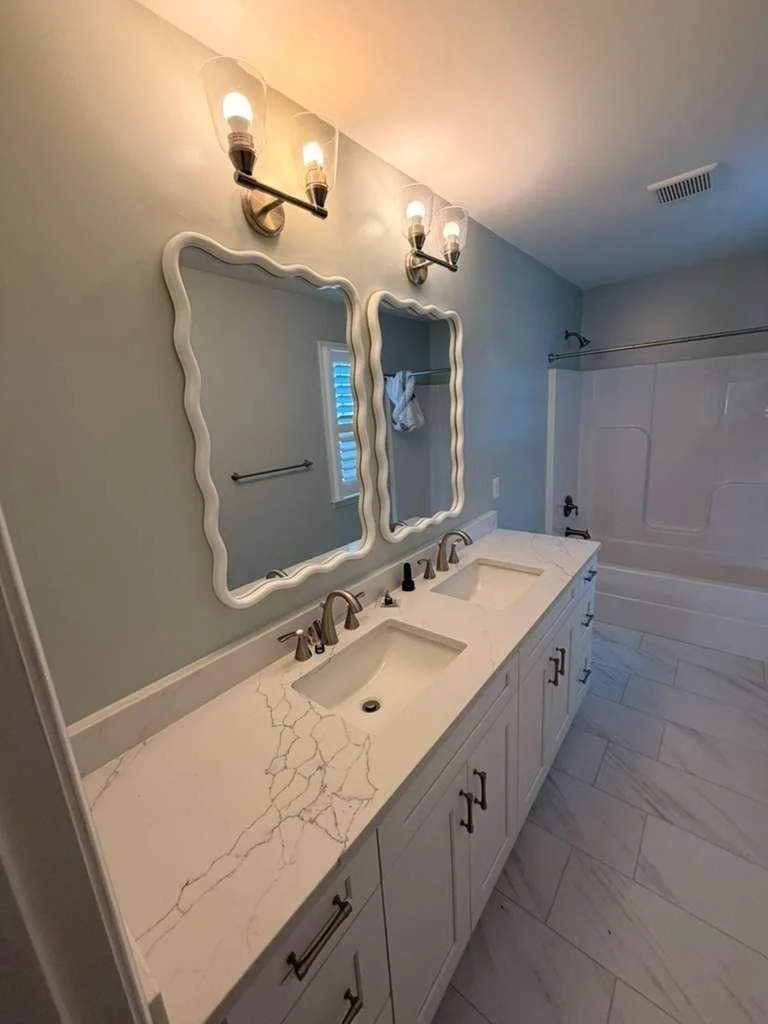 Bathroom with double vanity, white marble countertop with gray veining, two rectangular undermount sinks, two wavy-edged mirrors, and a bathtub with a shower curtain. The walls are painted light blue, and the floor is tiled with white marble tiles. There are two light fixtures with three exposed bulbs above the mirrors.