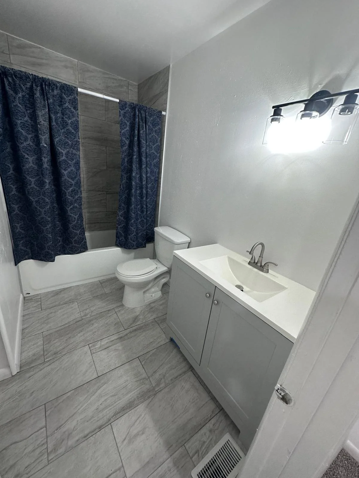 Small bathroom with white wall, gray tile flooring, a toilet, a small gray vanity with a sink and chrome faucet, a light fixture with three bulbs, and a bathtub with dark blue patterned shower curtains.