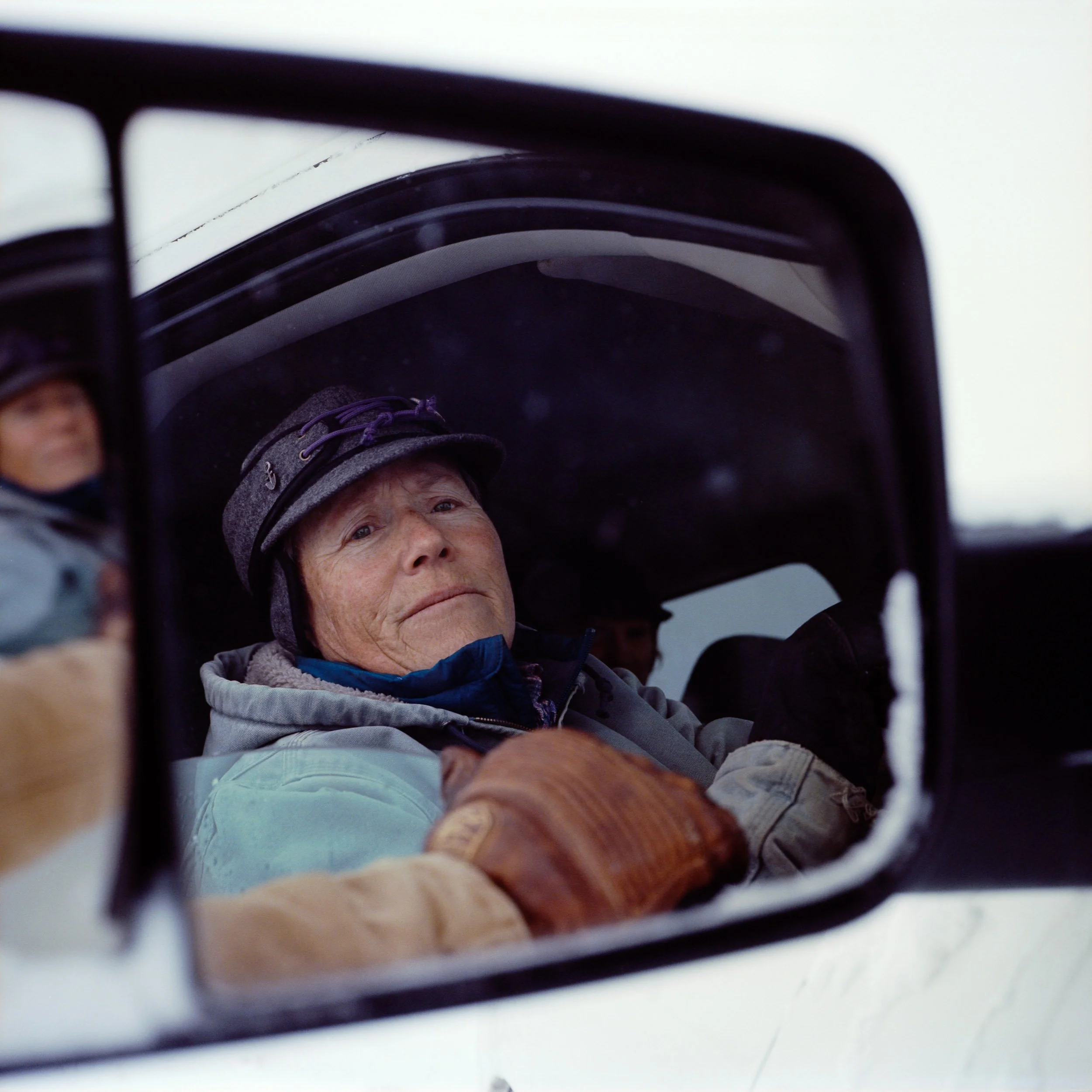 A woman looking into a vehicle's side mirror, with a reflection of her face, wearing a gray hat and glove, and gently holding a dog.